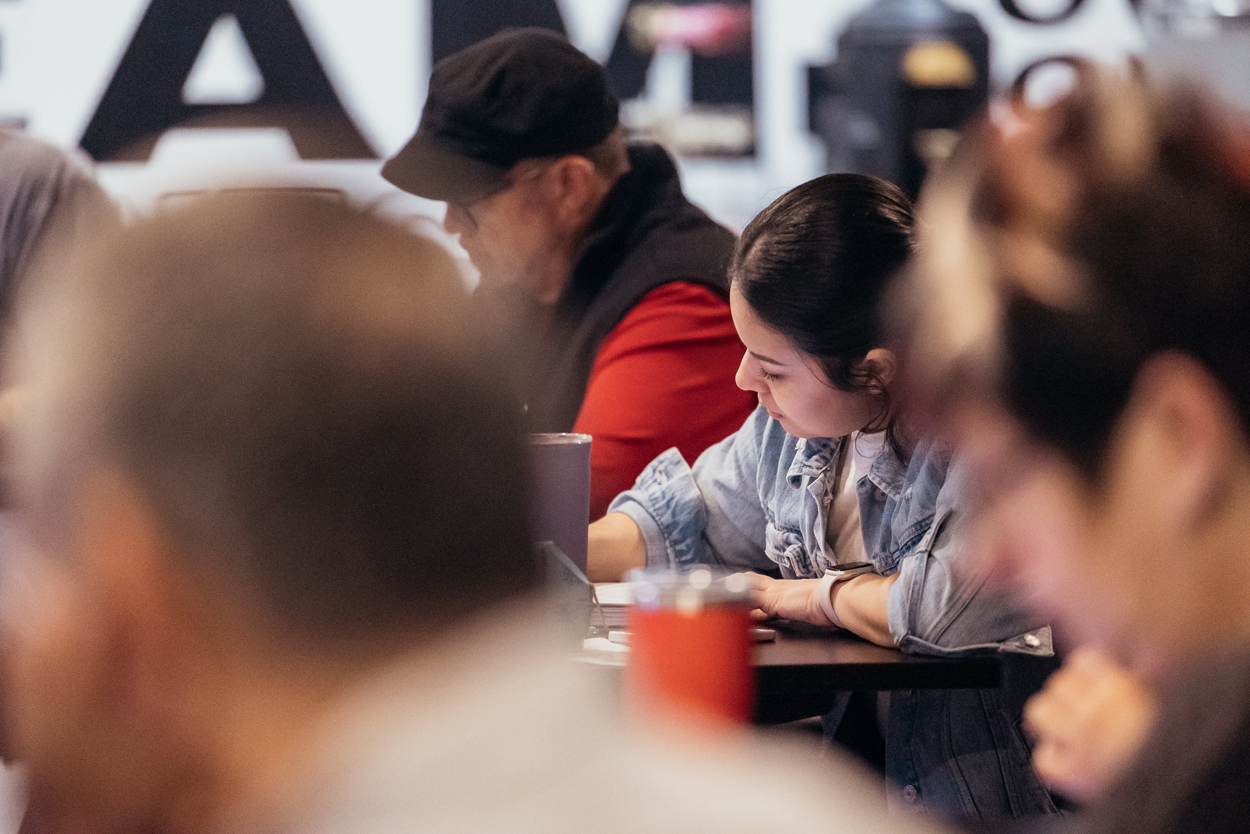 People sitting at a table in a café or restaurant, engaging in conversation and looking at their phones, with a large black letter 'A' in the background.