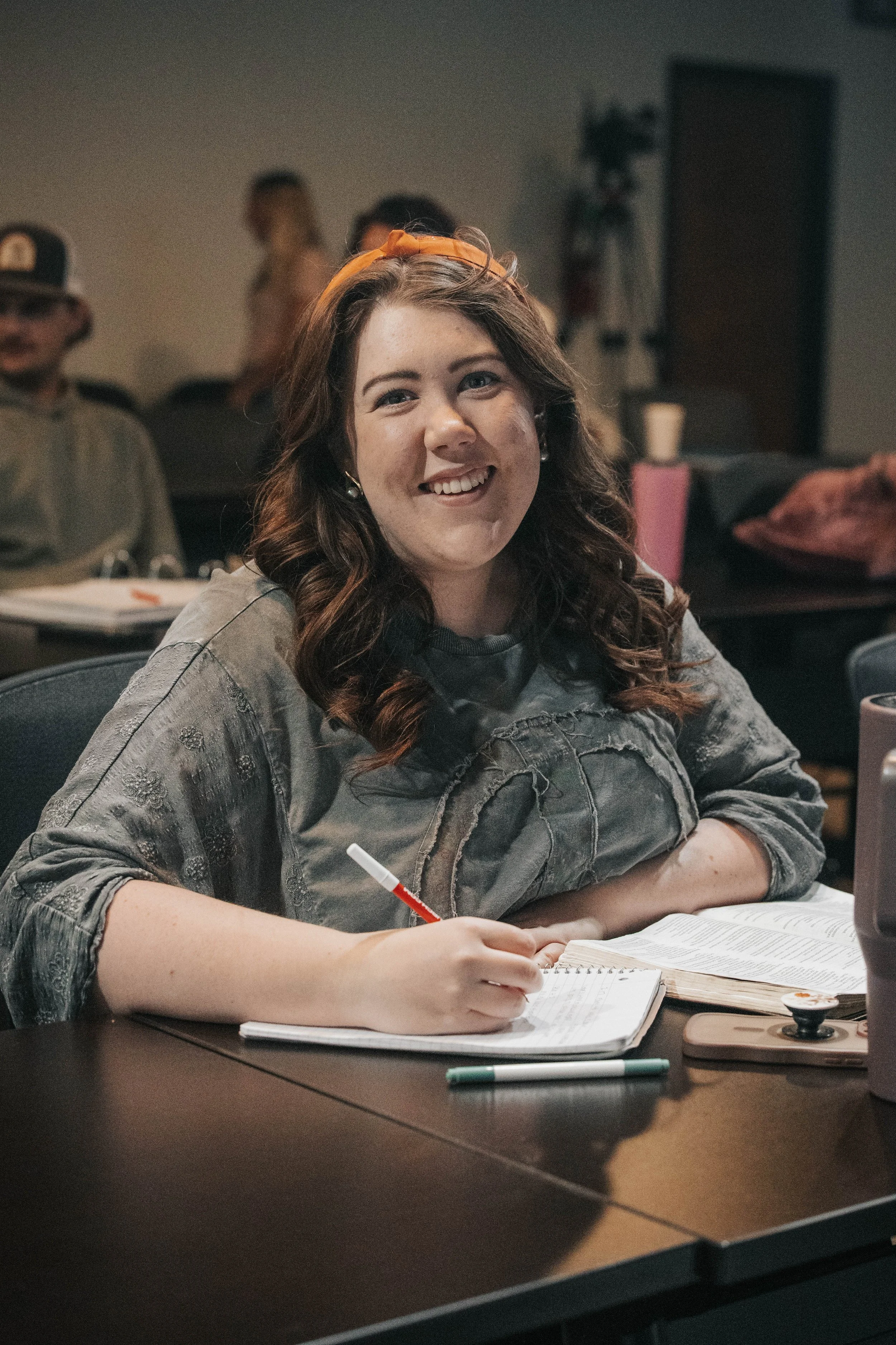 A young woman with brown hair and a headband, smiling and writing in a notebook at a table in a room with other people in the background.