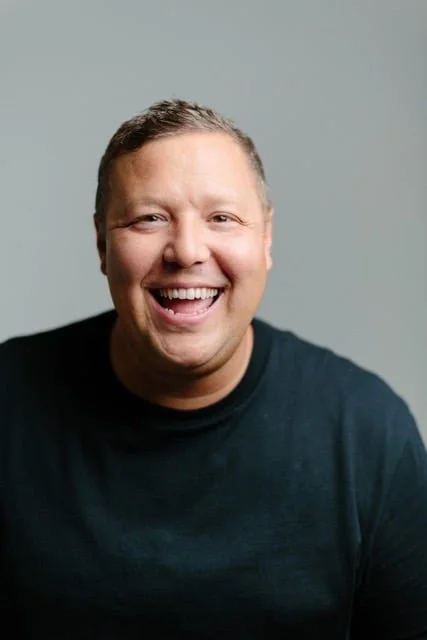 A man with short hair smiling and wearing a black shirt against a plain light gray background.