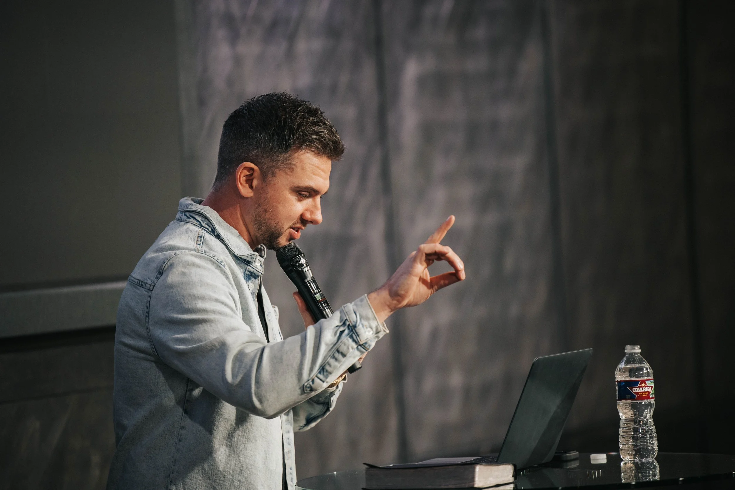 A man with short dark hair and beard, wearing a light denim jacket, speaking into a microphone while raising his right hand with one finger pointing upward. He is sitting at a table with a laptop and a bottle of Ozarka water.