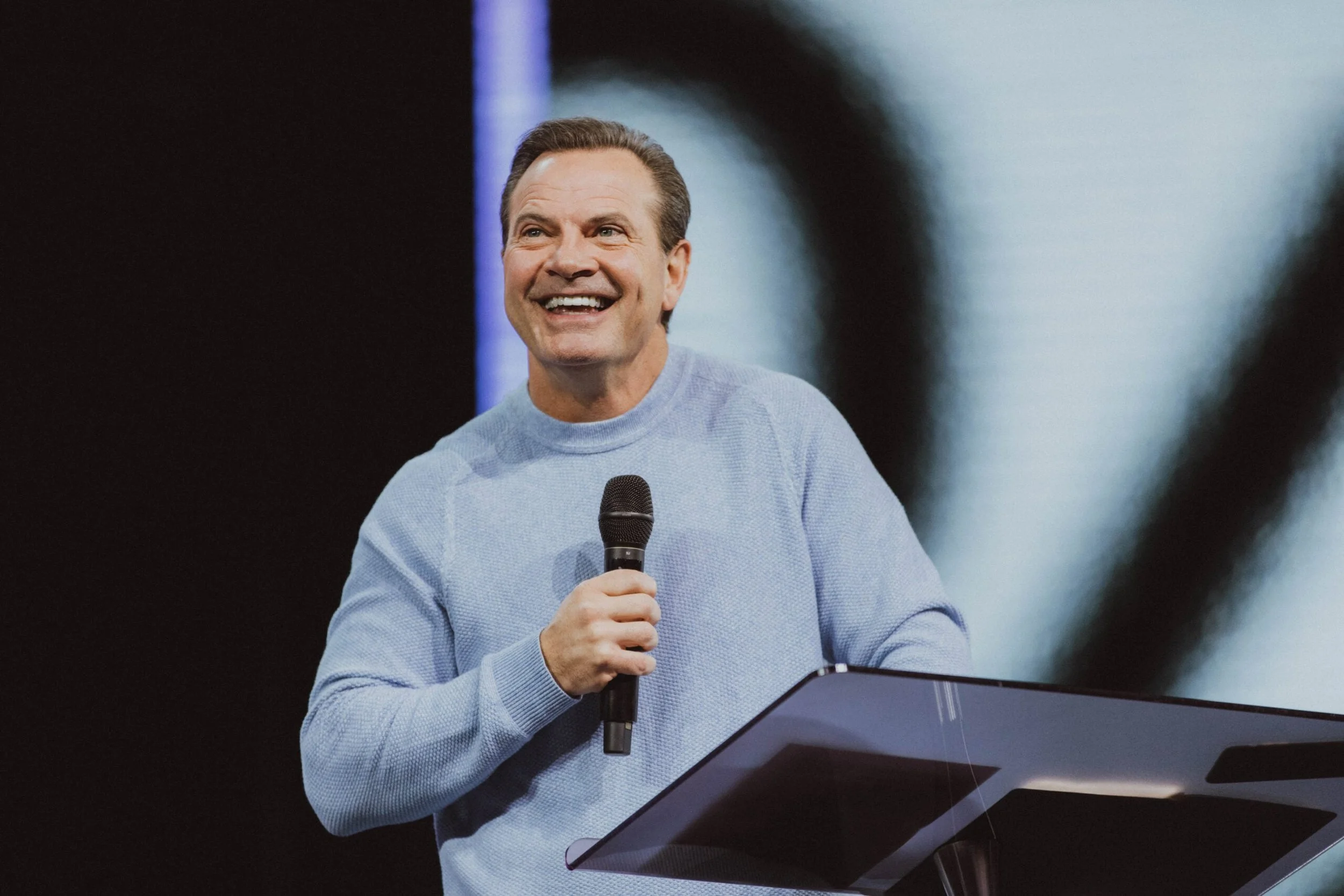 Man smiling and speaking into a microphone behind a lectern, wearing a light blue sweater.