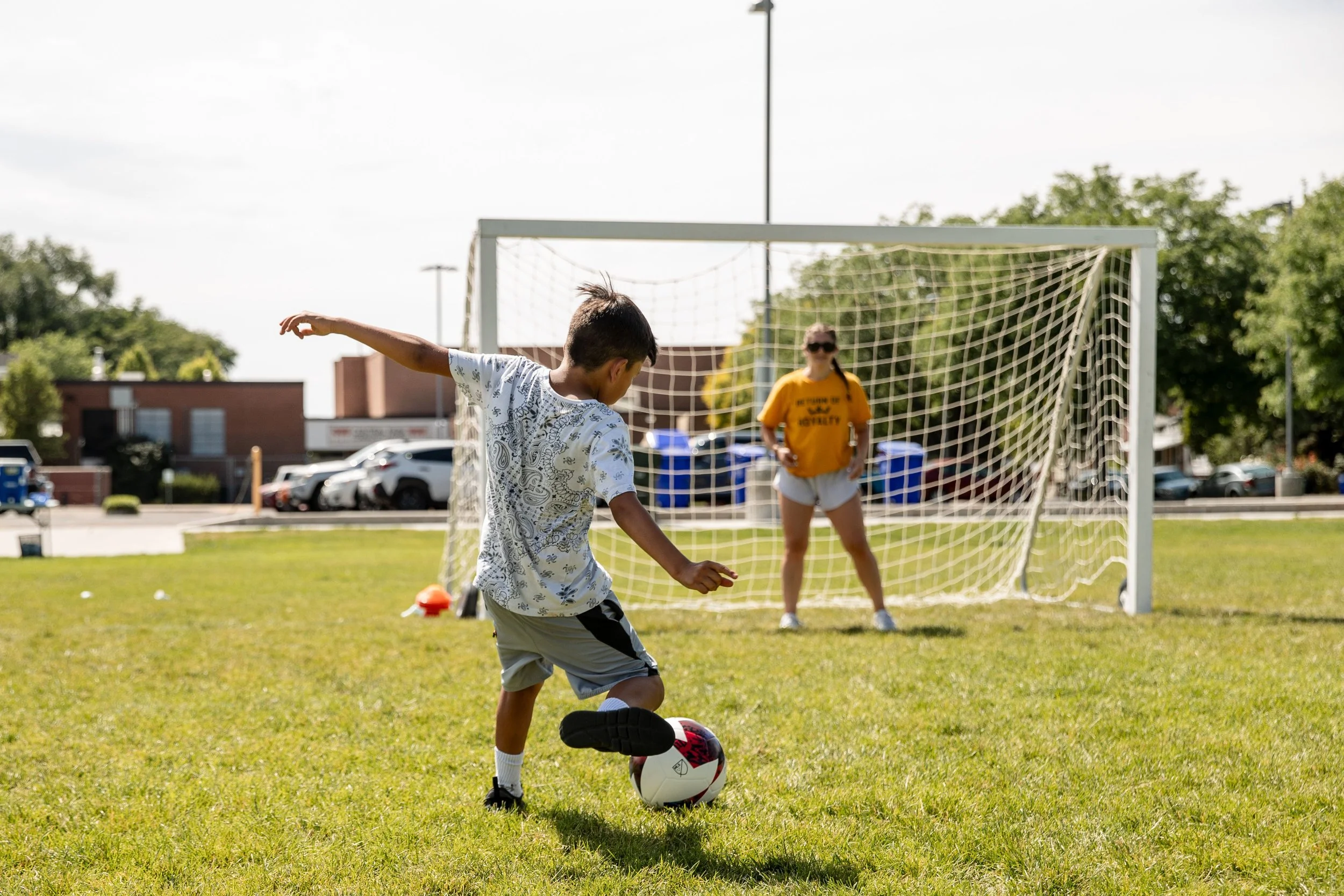 Kid kicking a soccer ball into a goal net with a goalie.