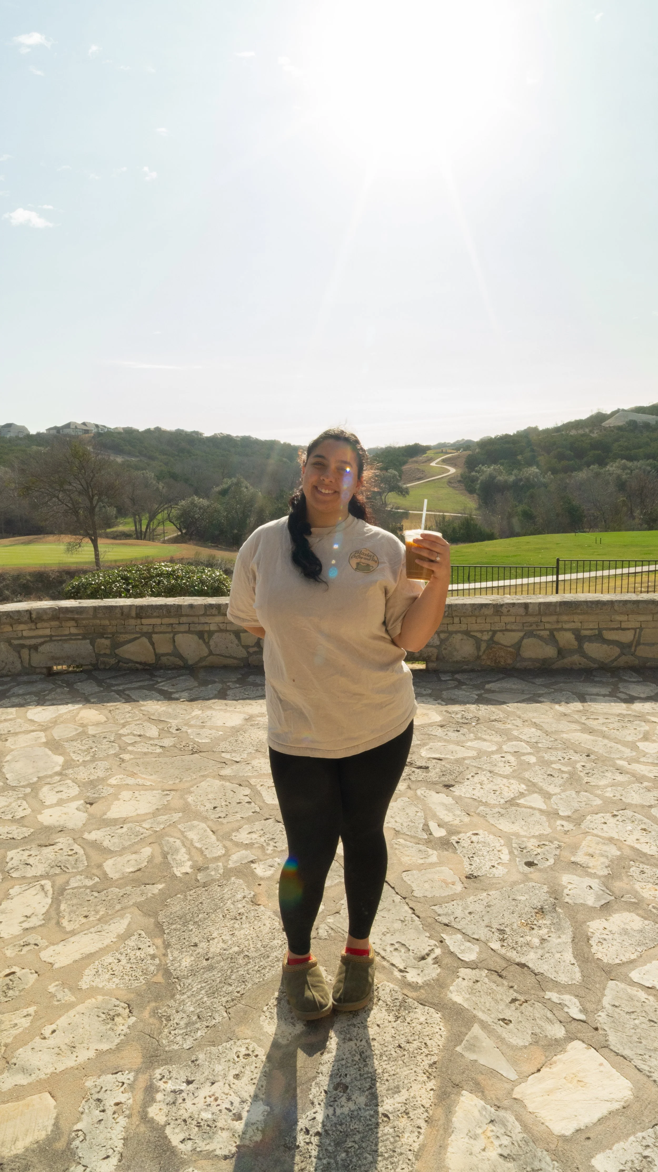 A woman smiling and holding a drink on a stone patio with a scenic view of rolling hills and a winding path in the background