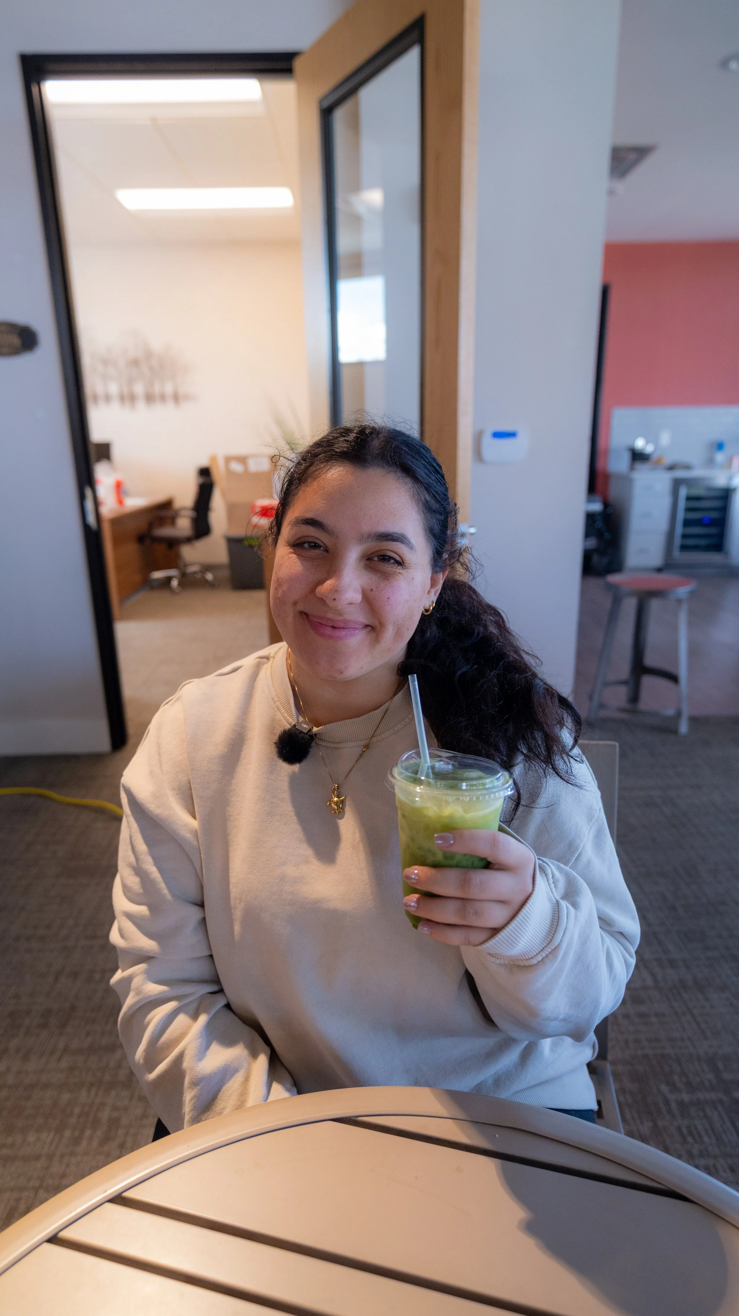 A young woman with dark, curly hair, smiling and holding a green smoothie with a straw, sitting at a table in an indoor space that appears to be an office break room.