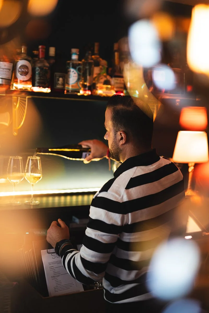 Ein Mann mit dunklem Haar und Bart, der eine Männerhand in einem schwarzen und weißen gestreiften Hemd, an einem Barhocker sitzend, Wein einschenkt, im Hintergrund befinden sich auf Regalen Flaschen und Gläser, mit warmem Beleuchtung und Lichter im H