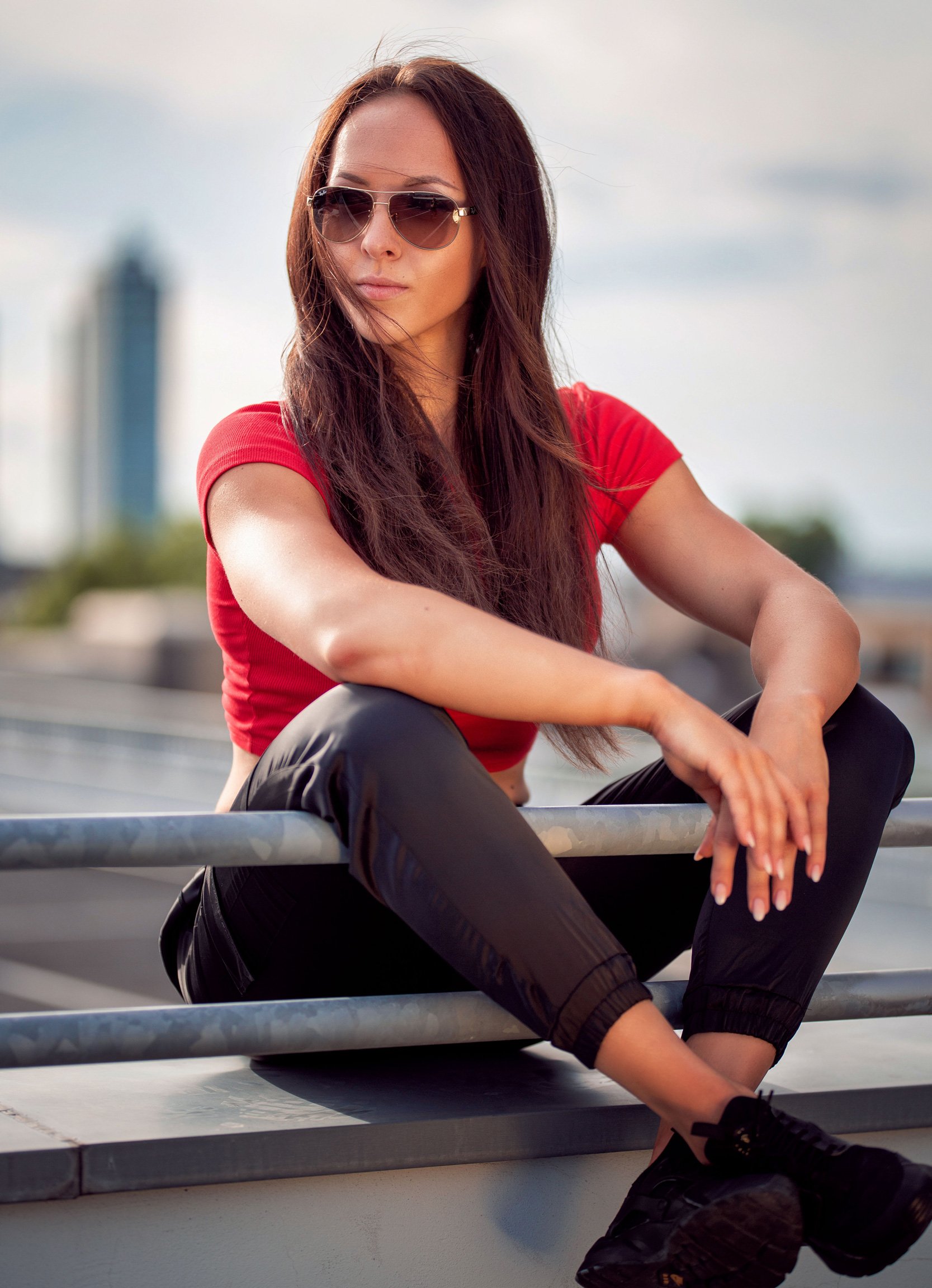 Junge Frau mit langen braunen Haaren, dunklen Sonnenbrillen, rotem Top, schwarzen Hosen und Sneakers sitzt auf einem Parkdeck, im Hintergrund Wolken und ein Stadtbild.
