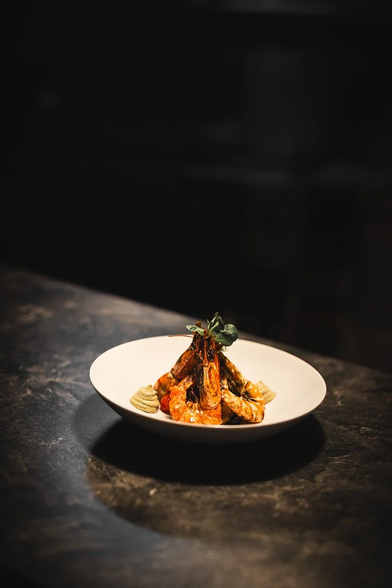 Ein weißer Teller mit Garnelen, garniert mit einem Zweig Grünzeug, auf einer dunklen Arbeitsplatte in einem schwach beleuchteten Raum.