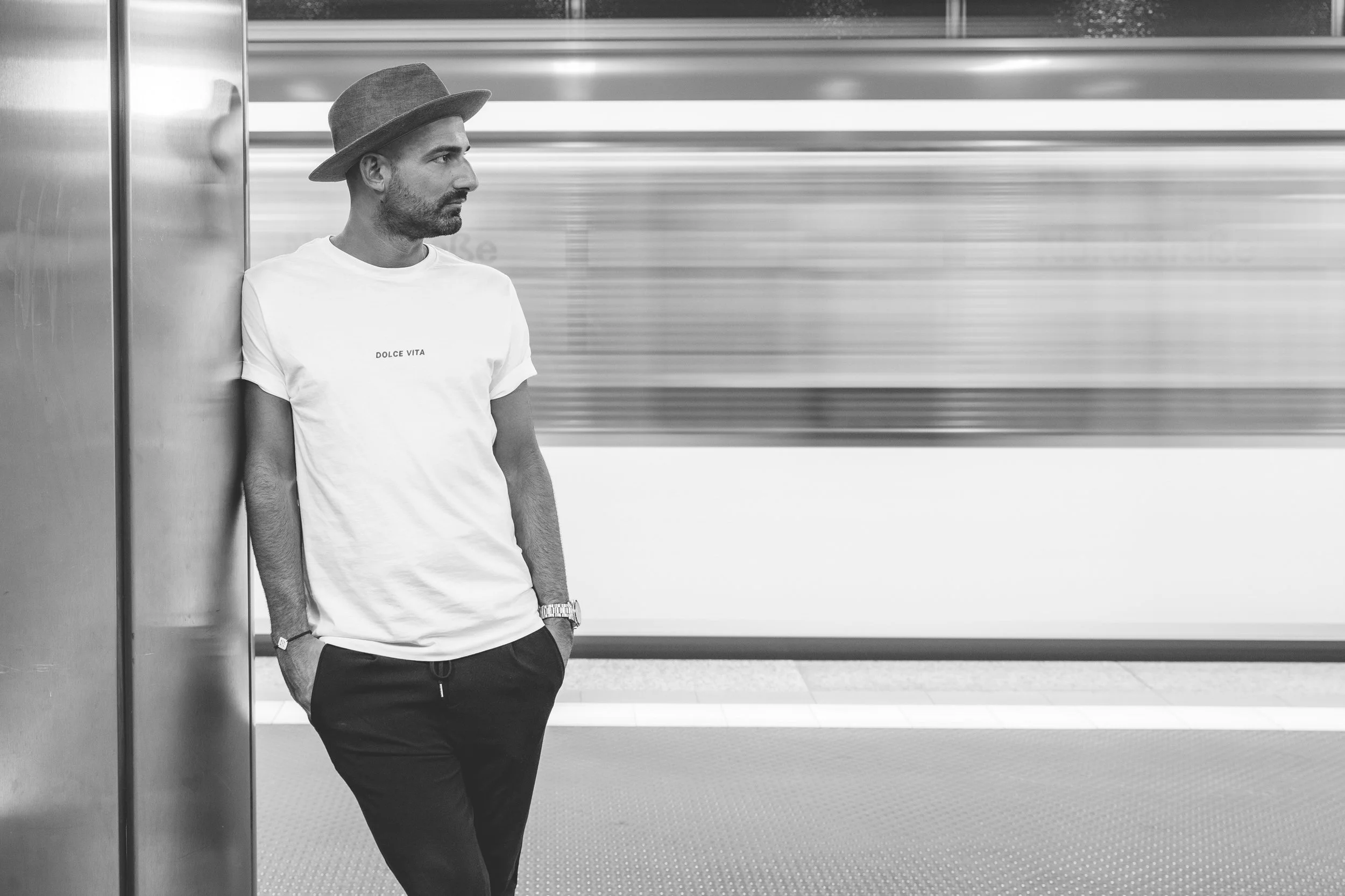 Mann steht in einem U-Bahnhof, lehnt an einer Wand, trägt einen Hut, T-Shirt mit der Aufschrift 'DOLCE VITA' und eine Armbanduhr, im Hintergrund ist ein Schnellzug im Bewegungsschein.