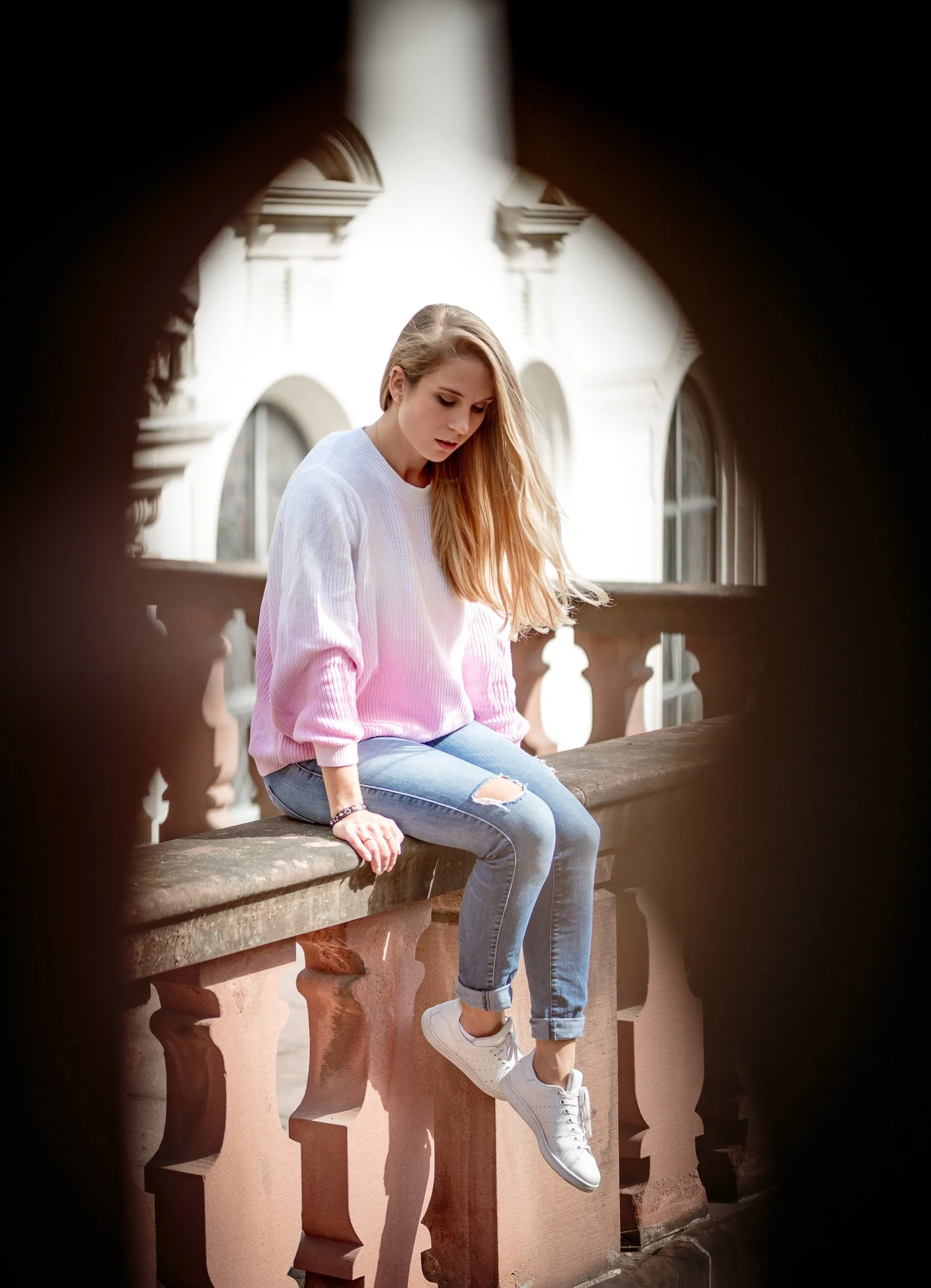Junge Frau in weißem Pullover, Jeans und weißen Sneakers sitzt auf einem Balkon, fotografiert durch ein Loch, vor einem historischen Gebäude.