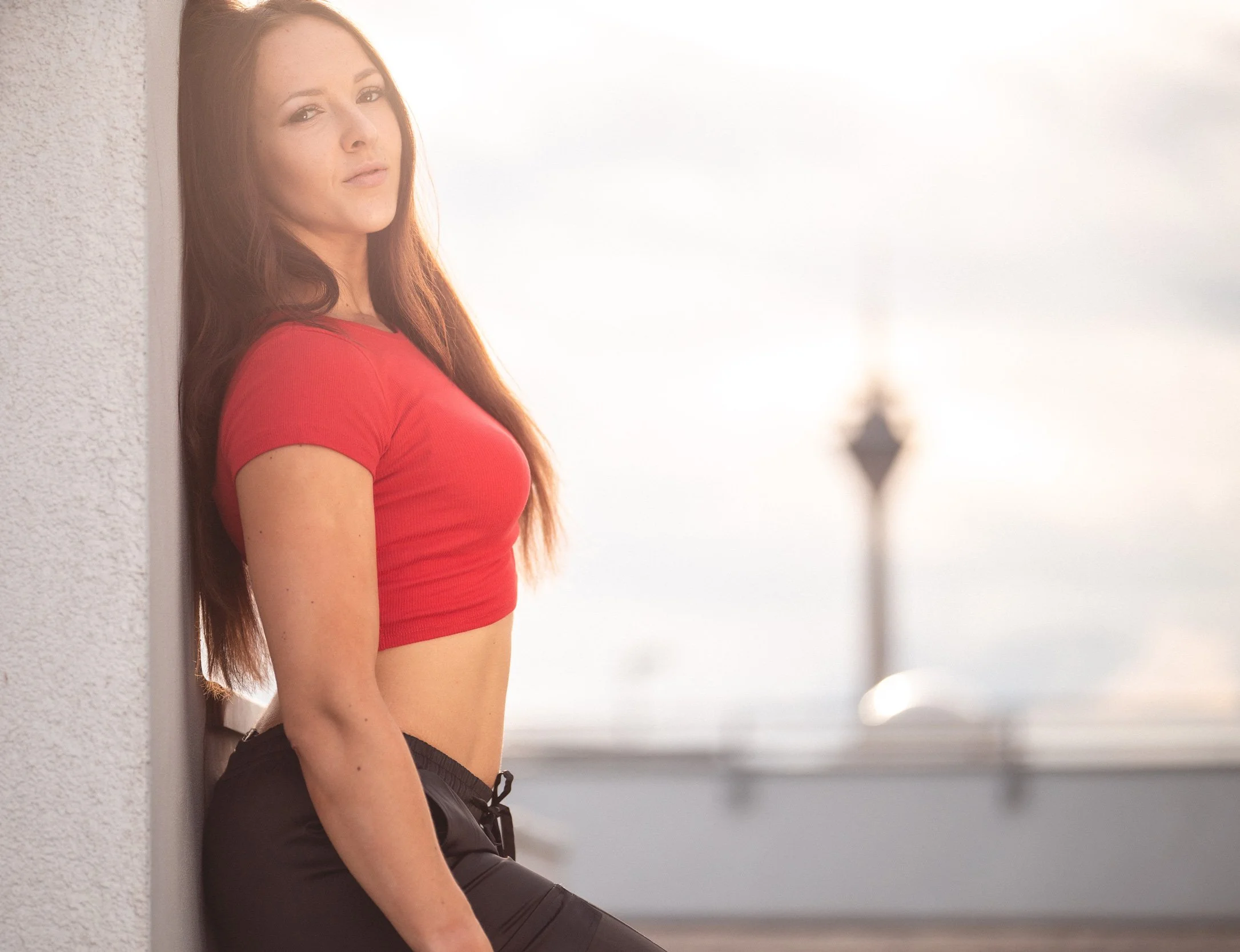 Eine Frau mit langen braunen Haaren, trägt ein rotes Crop-Top und schwarze Hosen, lehnt an einer weißen Wand im Freien, mit einem unscharfen Hochhaus im Hintergrund.