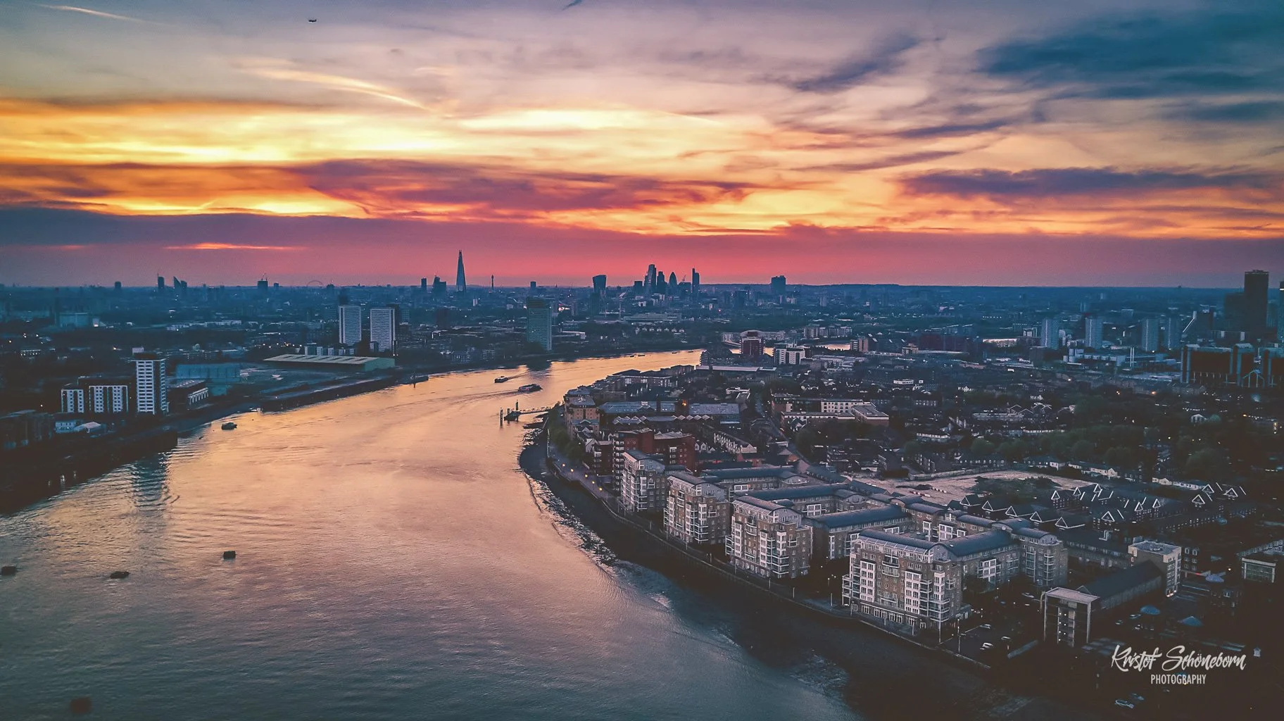 Stadtaufnahme von London bei Sonnenuntergang mit Fluss im Vordergrund und Wolken am Himmel, Skyline im Hintergrund.
