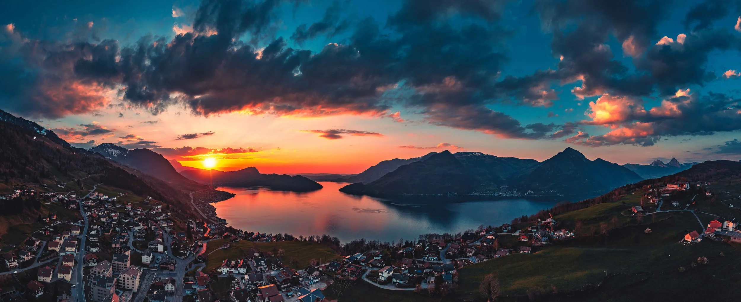 Blick auf einen Sonnenuntergang über einem See, umgeben von Bergen und einer kleinen Stadt mit Häusern und Straßen.