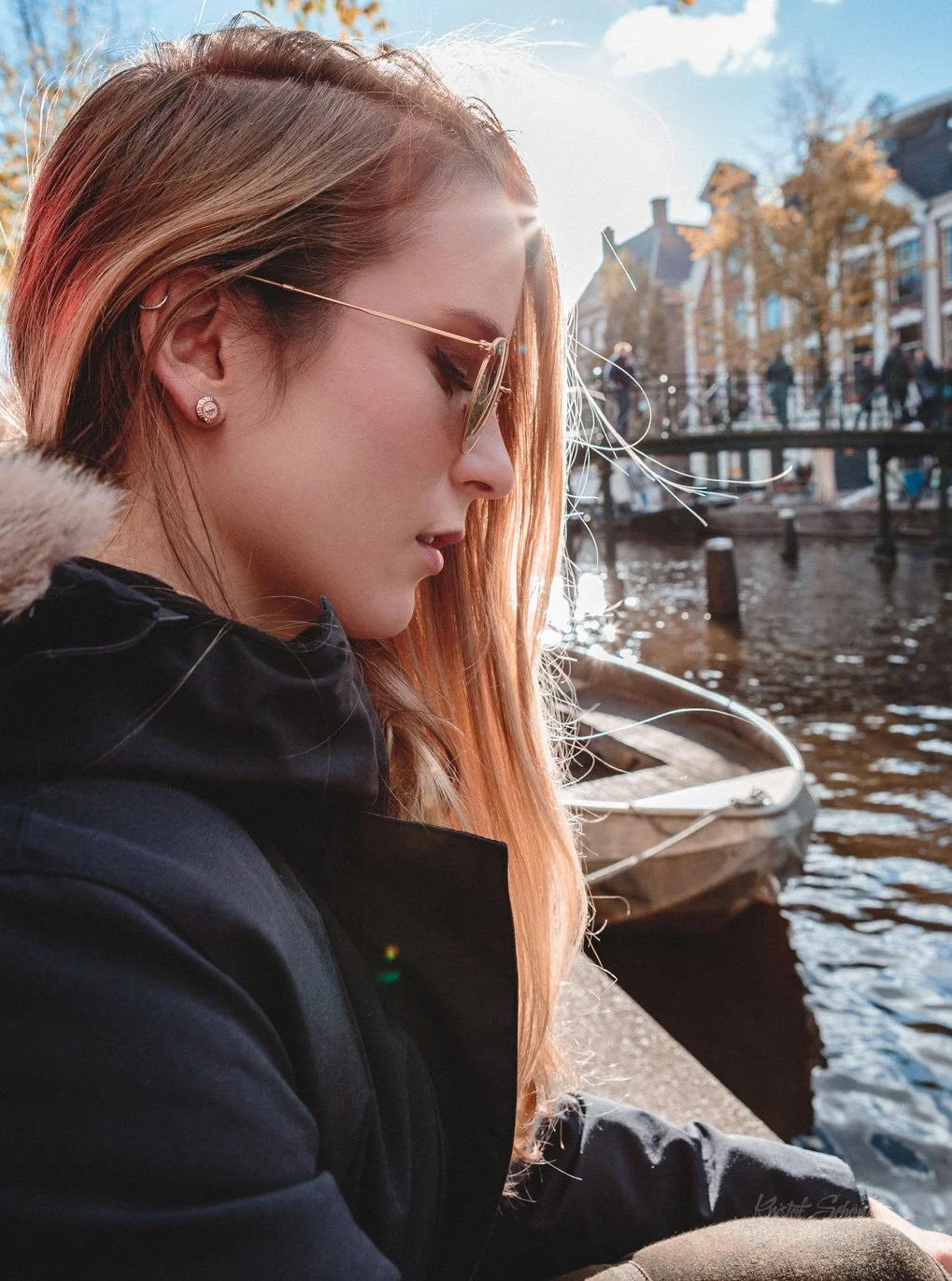 Seitliche Aufnahme einer jungen Frau mit langen, roten Haaren, Sonnenbrille und Ohrringen, die an einem Kanal in einer Stadt sitzt, mit einem Boot im Wasser und Gebäuden im Hintergrund, bei sonnigem Wetter.