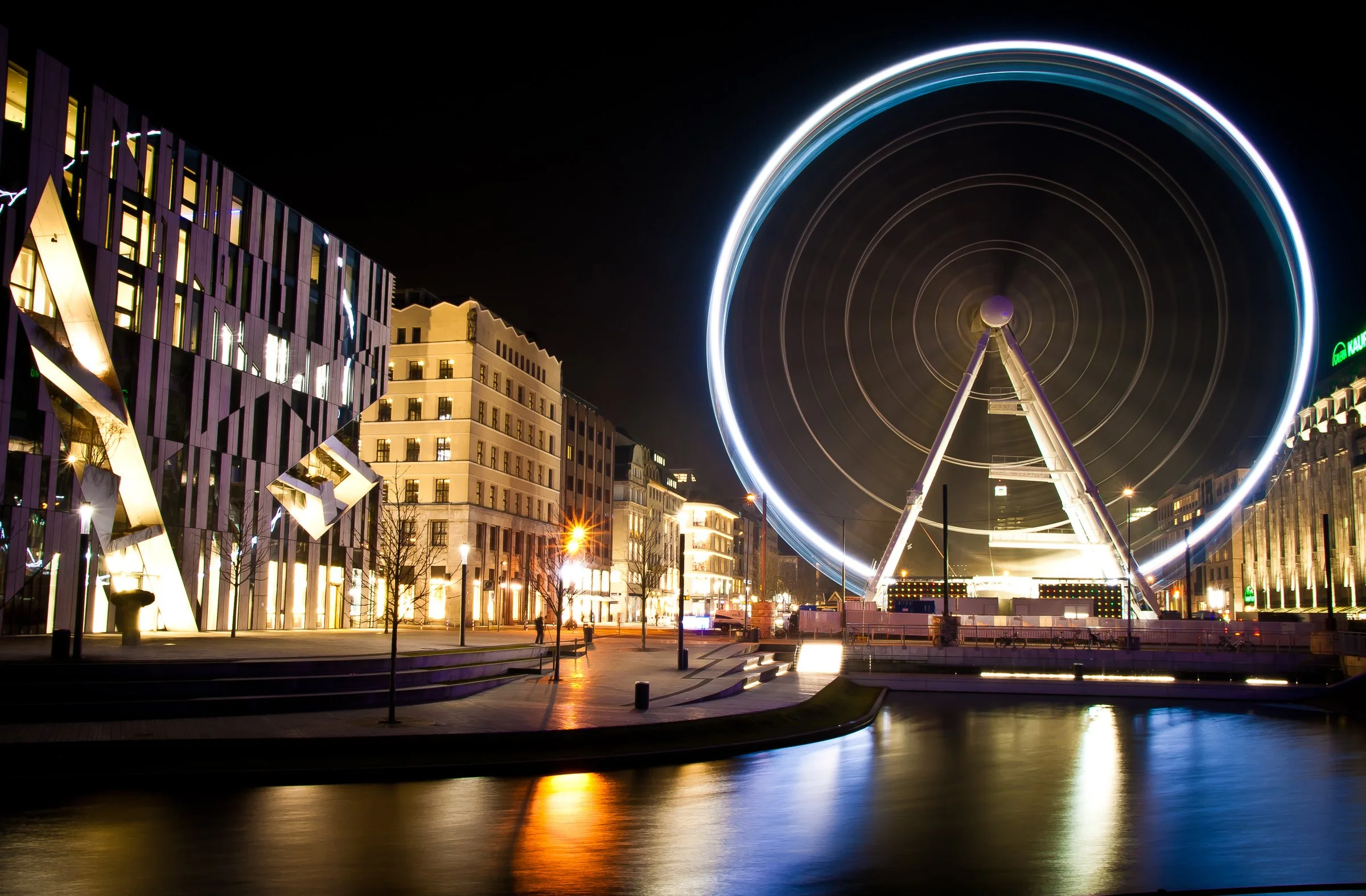Stadtbild bei Nacht von Düsseldorf mit beleuchtetem Riesenrad, modernen Gebäuden und Wasser im Vordergrund, das die Lichter reflektiert.