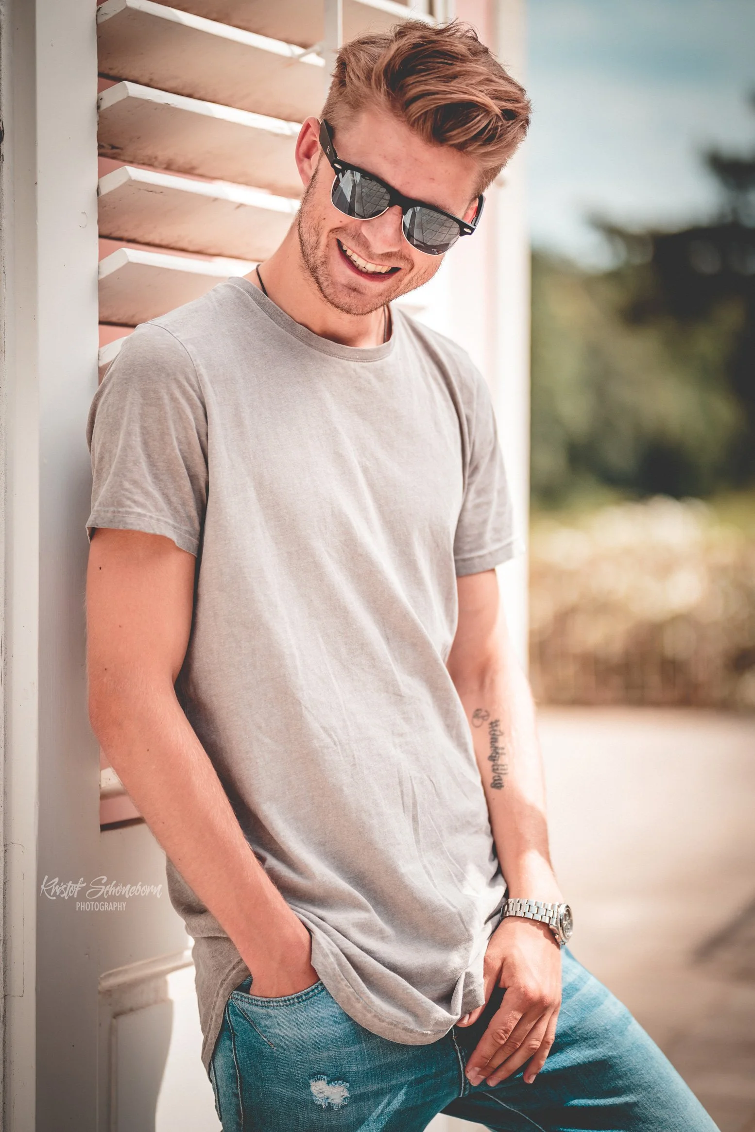 Lächelnder junger Mann mit Sonnenbrille, graues T-Shirt und Jeans, steht draußen vor einem Gebäude.