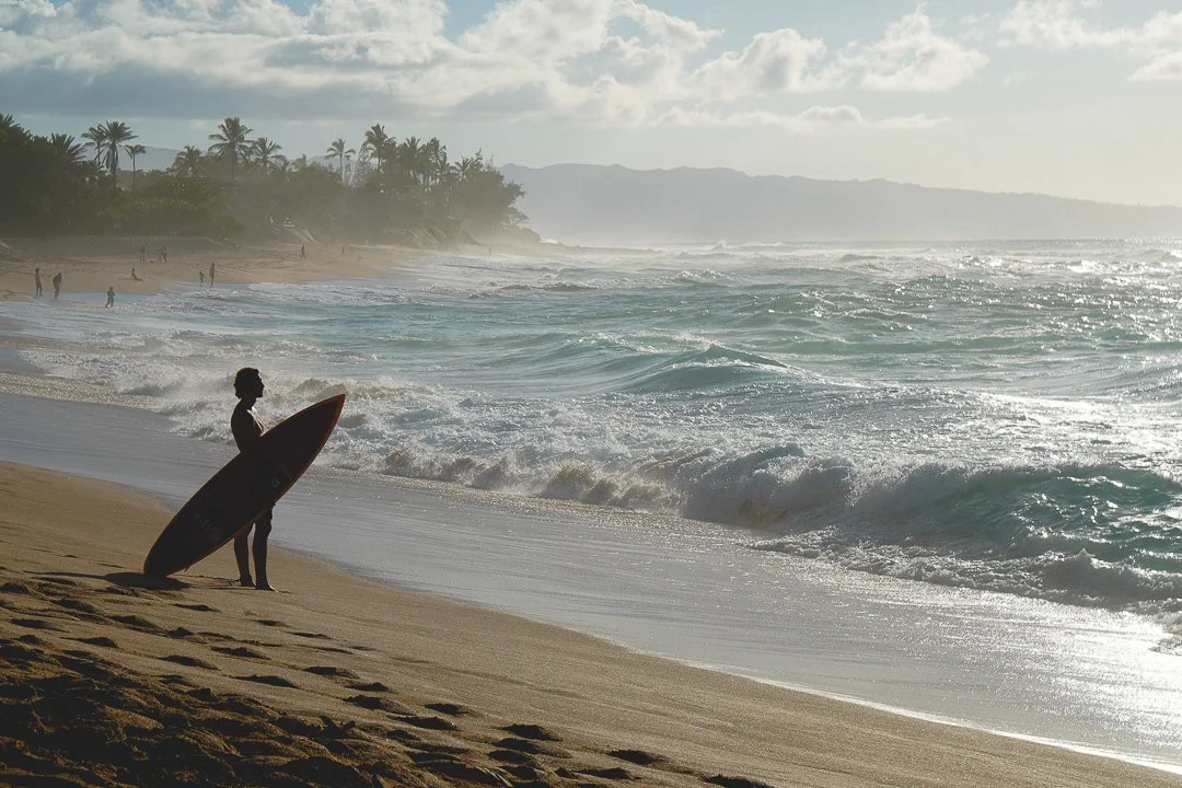 Ein Surfer mit Surfbrett am Strand bei Sonnenuntergang, Palmen im Hintergrund, Wellen im Meerwasser