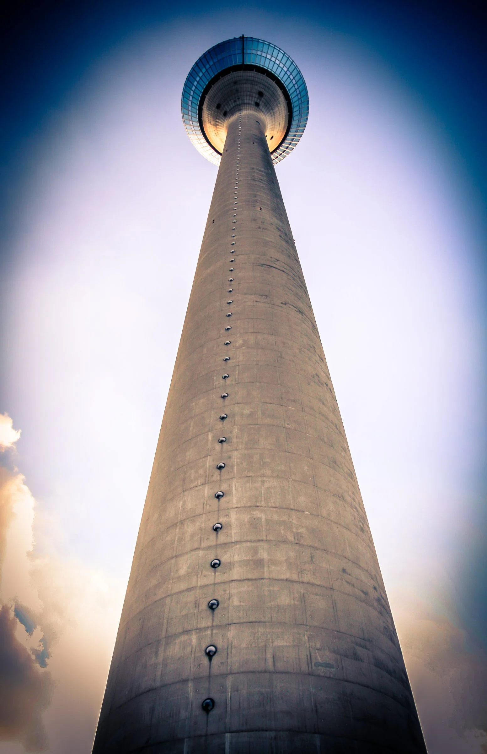 Blick nach oben auf den Fernsehturm von Düsseldorf mit runder Aussichtsplattform an der Spitze, fotografiert bei Sonnenaufgang oder -untergang, mit sichtbaren Sonnenstrahlen im Hintergrund.