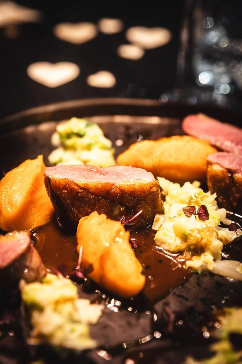 Nahaufnahme von gebratenem Steak, Kartoffeln, Brokkoli und Kartoffelpüree auf einem schwarzen Teller vor einem unscharfen Hintergrund aus herzförmigen Lichtern.