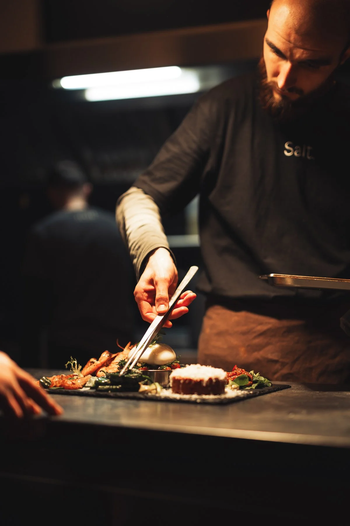 A chef in a black shirt with the word 'Salt' on it is plating food in a dimly lit kitchen. There are various dishes including a piece of cake and garnishes on the table.