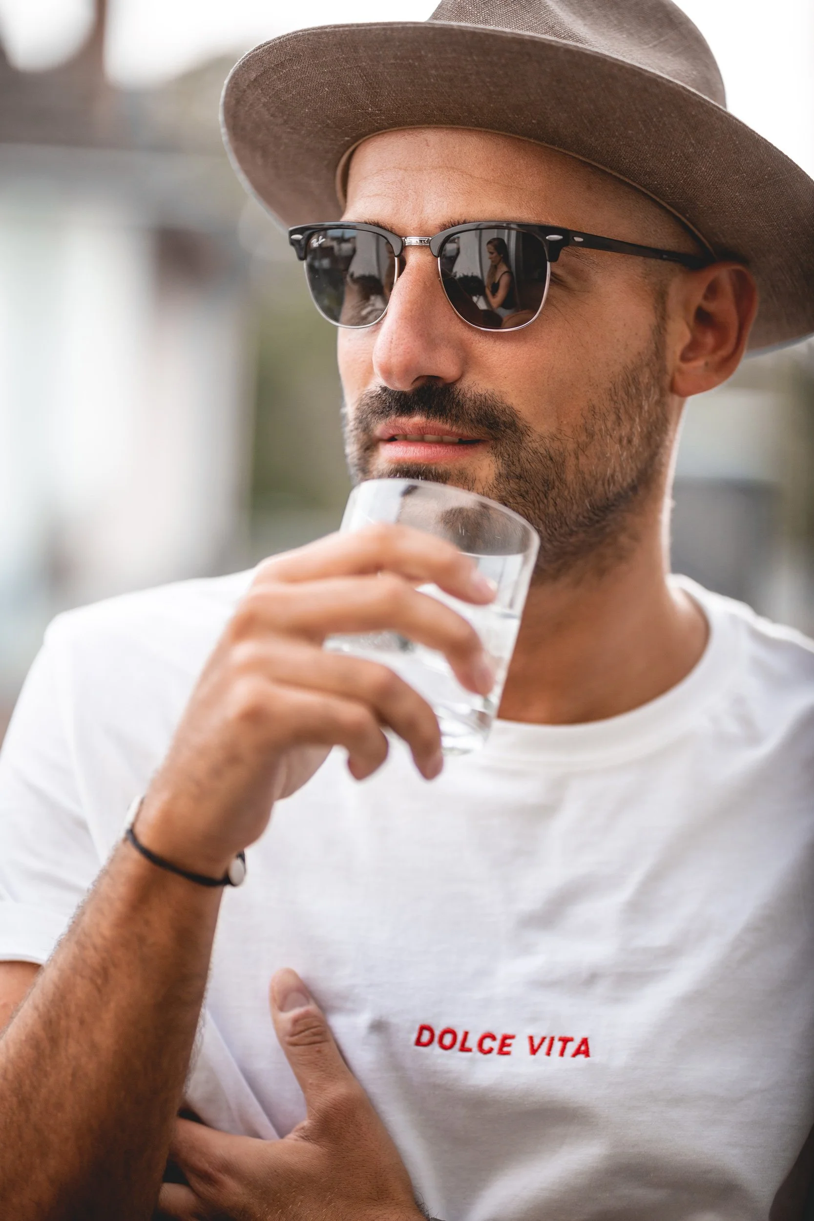 Ein Mann trägt einen Hut und Sonnenbrille, hält ein Glas Wasser und trägt ein weißes T-Shirt mit rotem Schriftzug 'DOLCE VITA'.