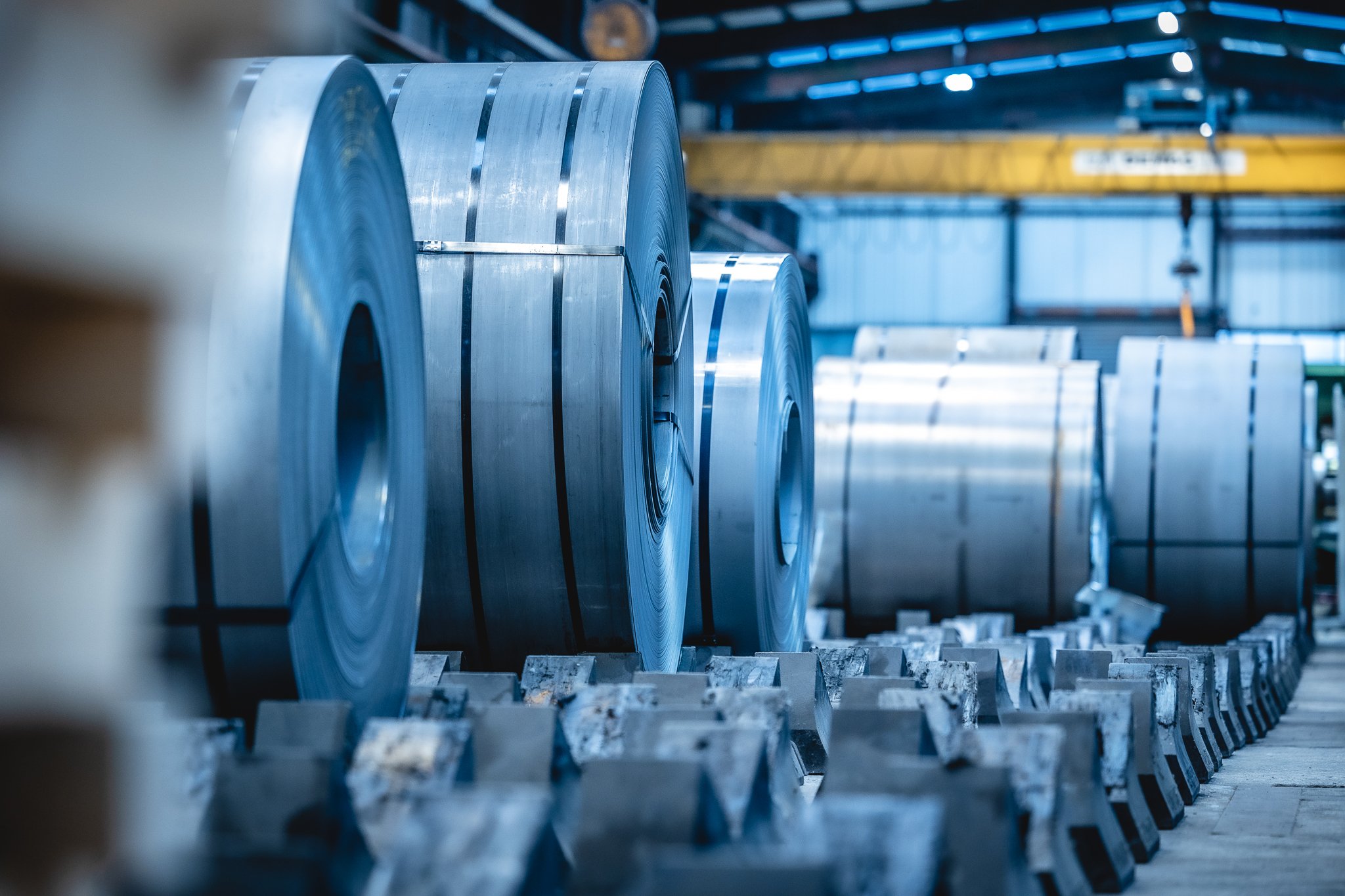 Große Stahlcoils, die auf Stützen in einer Fabrik oder einem Lagerhaus ruhen, im Hintergrund ein gelber Laufkran.