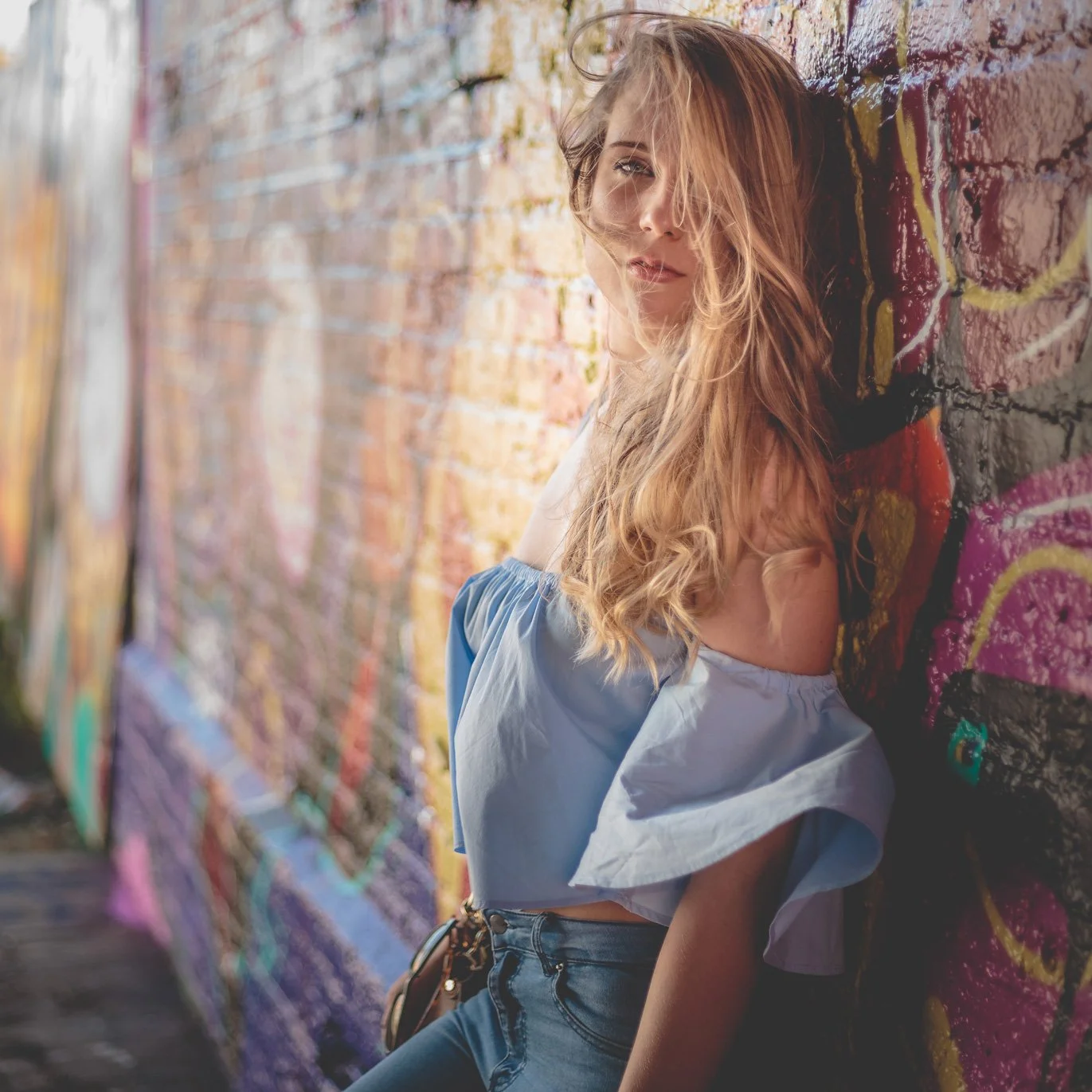 Junge Frau mit langem, lockigem Haar lehnt an einer mit Graffiti bemalten Wand, im Hintergrund eine bunte Graffiti-Wand.