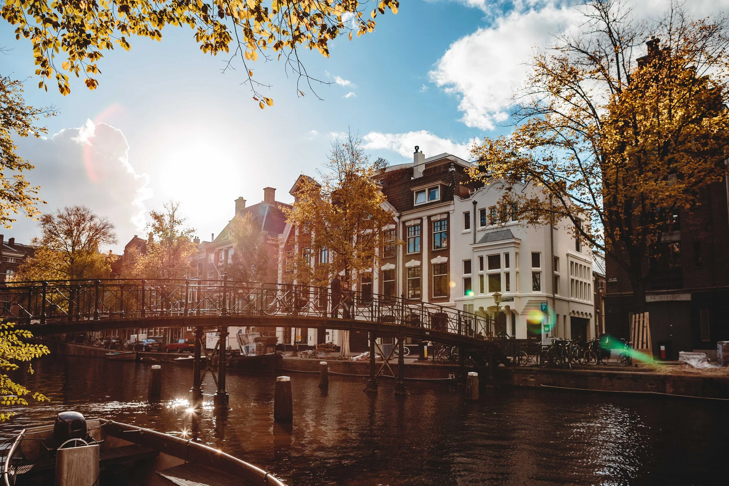 Blick auf eine Brücke über einen Kanal in einer Stadt im Sonnenschein, mit fahrrädern und historischen Gebäuden im Hintergrund, umgeben von herbstlichen Bäumen.