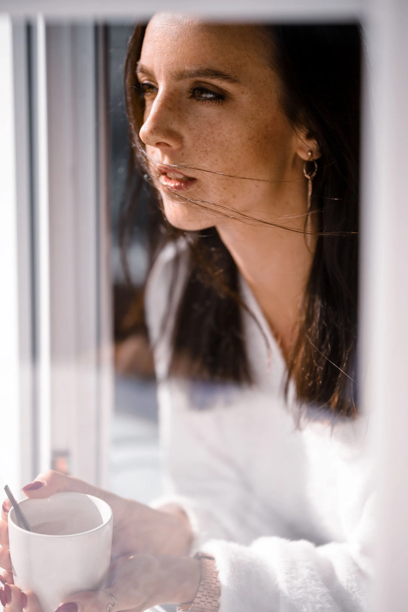 Frau mit dunklen Haaren, der in eine Fensterscheibe schaut, hält eine weiße Tasse in der Hand.