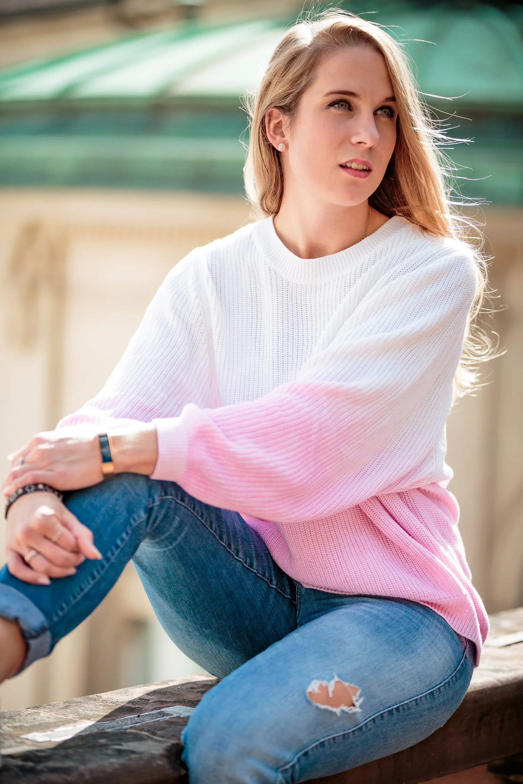 Junge Frau mit hellblondem Haar in Weiß und Pink sitzend auf einem Holzblock, blickt nach rechts, im Sonnenschein, im Freien.