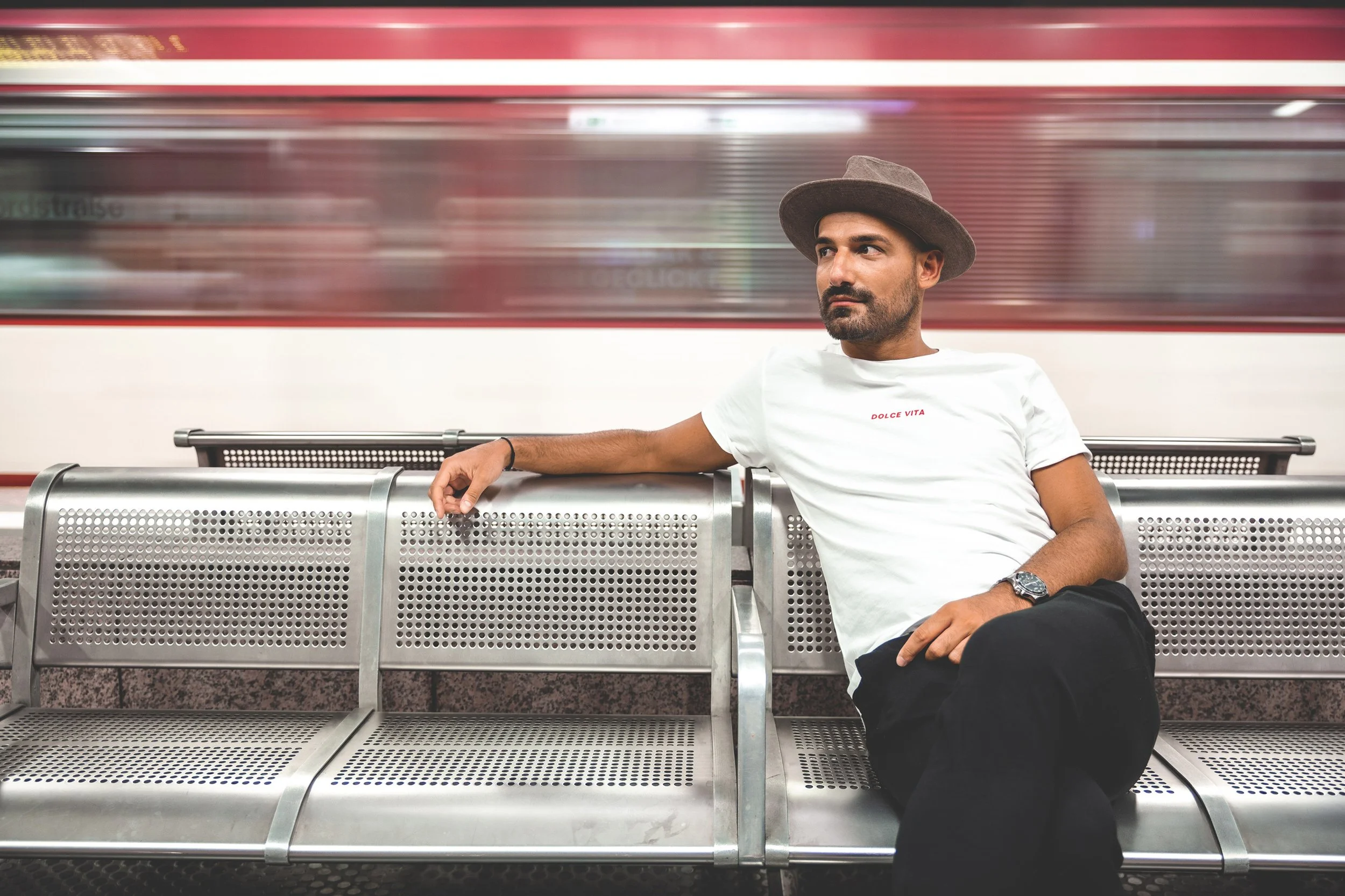 Ein Mann sitzt auf einer Bank in einer U-Bahn-Station, trägt einen Hut und ein weißes T-Shirt mit rotem Text, während die U-Bahn im Hintergrund vorbeifährt.