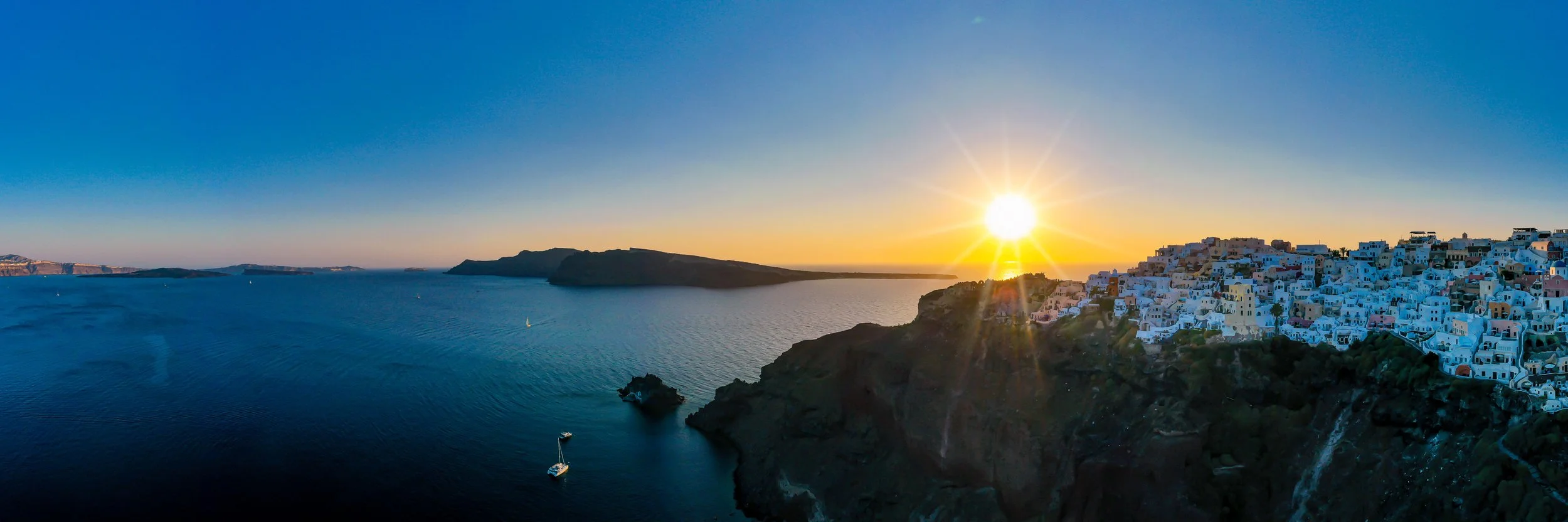 Blick auf das Meer bei Sonnenuntergang mit weißen Gebäuden auf einer Klippe auf der rechten Seite.