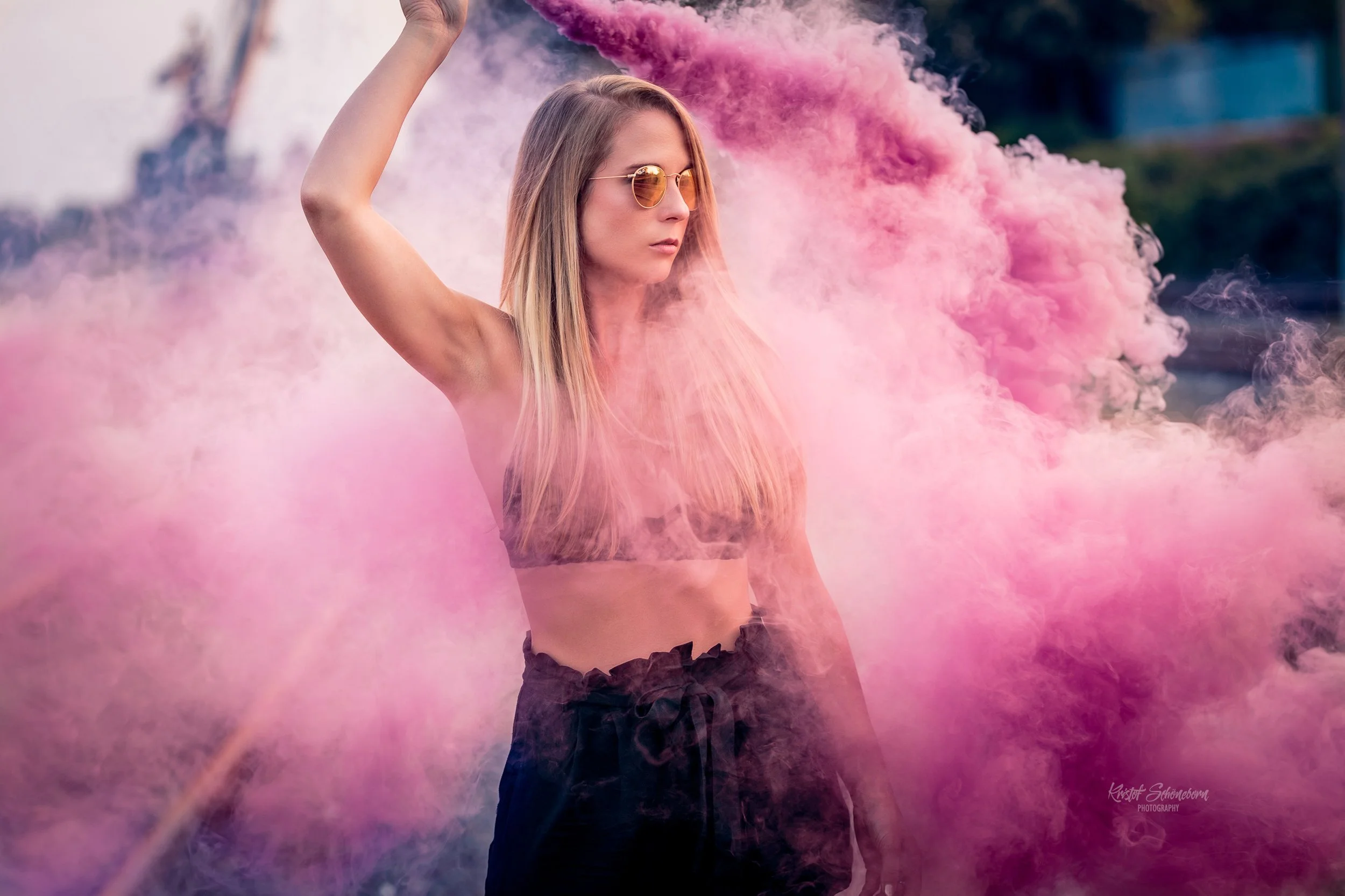 Eine junge Frau mit Sonnenbrille posiert inmitten von pinkem Rauch. Sie trägt schwarze Shorts und ein schwarzes Top. Der Hintergrund ist unscharf.