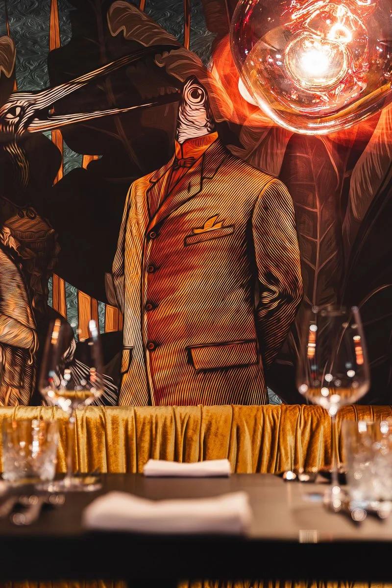 Ein stilisiertes Wandbild, das einen Mann im Anzug zeigt mit Fischkopf, vor dessen Gesicht eine große Glaskugel platziert ist, was einen surrealen Effekt erzeugt; im Vordergrund ist ein Esstisch zu sehen, auf dem Weingläser und Servietten stehen.