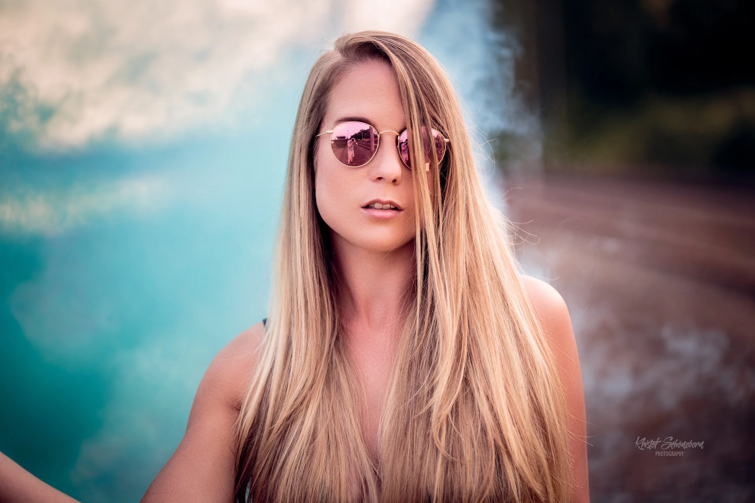 Junge Frau mit langen, blonden Haaren und rosa Sonnenbrille posiert vor einem Wasserfall im Freien.