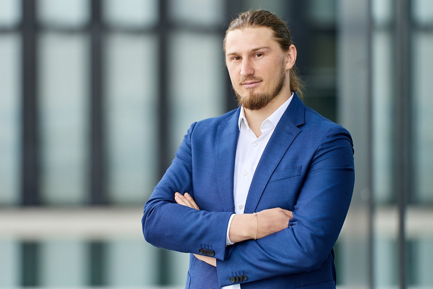 Business Portrait von einem jungen Mann in blauer Anzug mit verschränkten Armen, vor moderner Gebäudefassade.