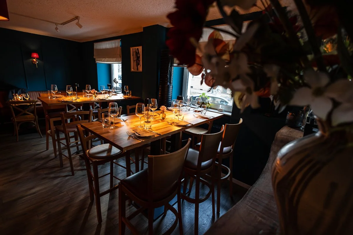 Eleganter, dunkel erleuchteter Essraum mit Wein- und WasserGläsern auf Tischen, großes Fenster mit Blick nach draußen, Blumen in Dekorvasen, blaue Wände und Holzfußboden.