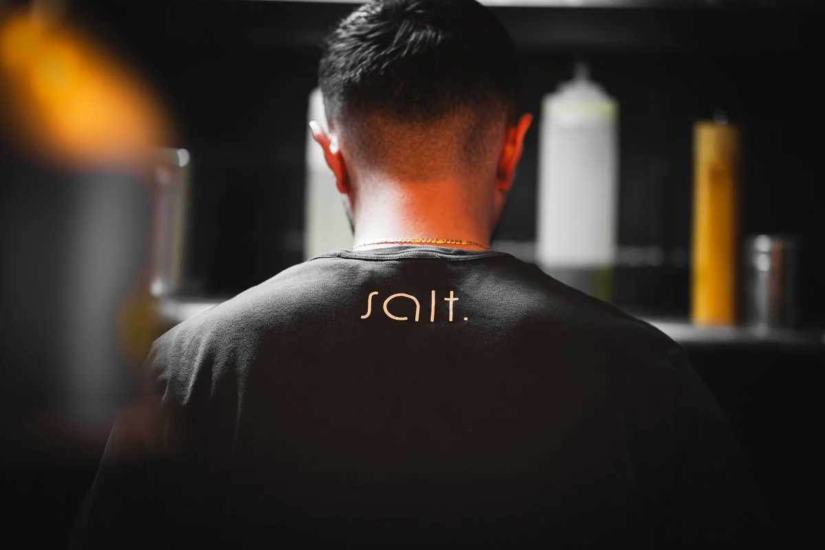 Der Rücken einer Person, die ein schwarzes T-Shirt mit der Aufschrift „Salt“ trägt, von hinten gesehen in einer schwach beleuchteten Umgebung mit Flaschen und Gewürzen im Hintergrund.