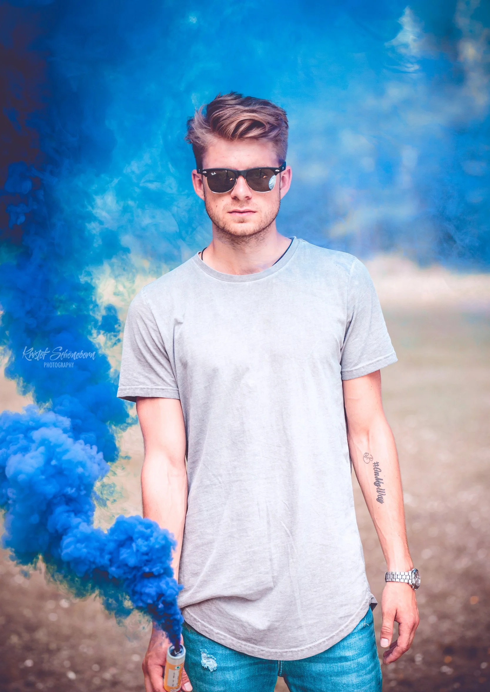 Junger Mann mit Sonnenbrille hält blaue Rauchbombe in der Hand, steht auf einer Wiese, trägt ein graues T-Shirt und blaue Jeans.