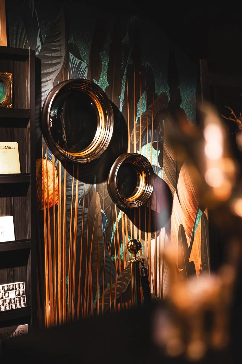 Eine dekorative Wand mit großen, glänzenden, schwarzen runden Spiegeln und einer botanischen Tapete mit großen tropischen Blättern und filigranen goldenen und grünen Akzenten.