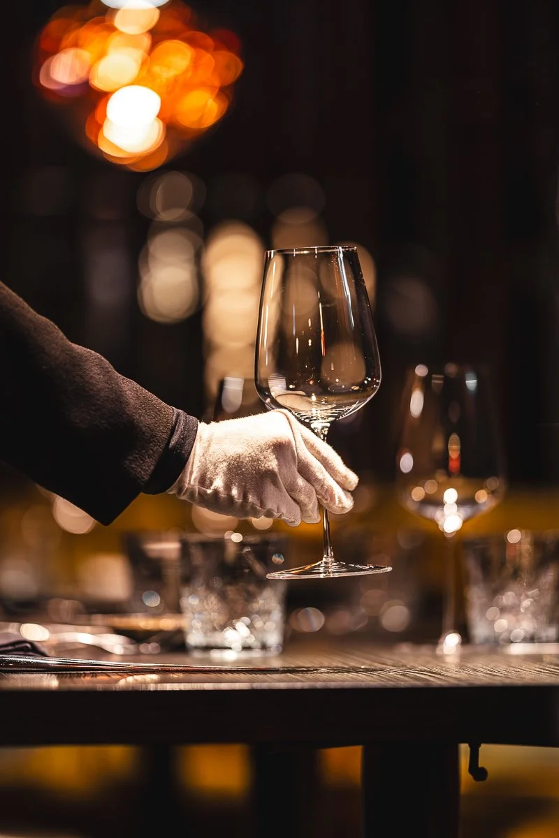 Eine Person in einem schwarzen Kleid trägt weiße Handschuhe und hält ein leeres Weinglas in einer handebenen Haltung. Der Hintergrund ist dunkel mit warmem, verschwommenem Licht, das eine festliche Atmosphäre schafft.