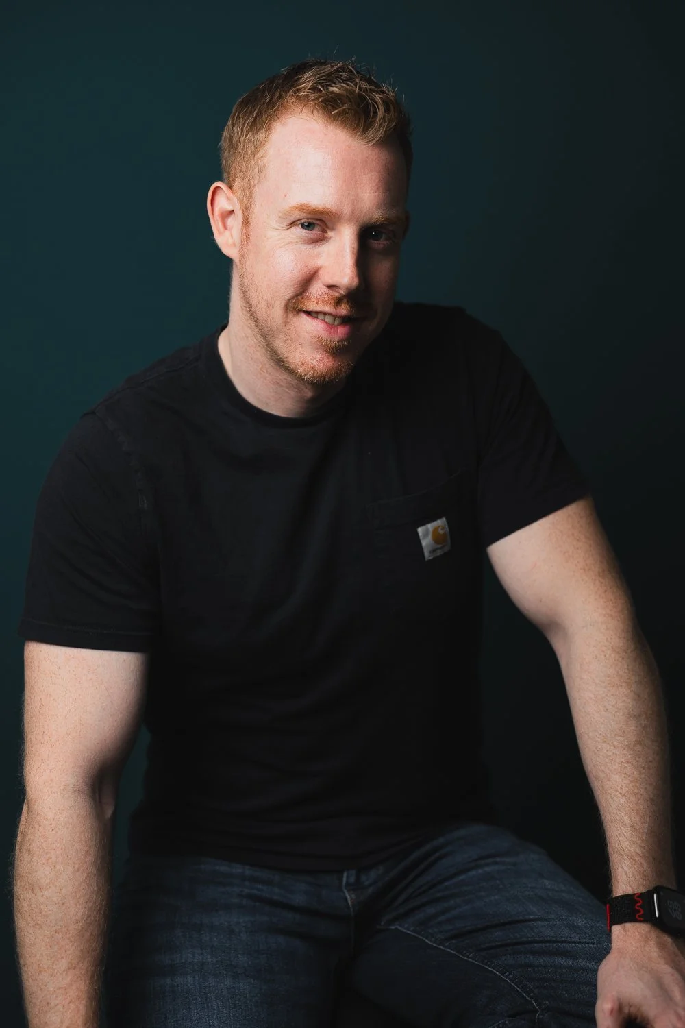 Mann mit roten Haaren und Bart, trägt schwarzes T-Shirt und sitzt vor dunklem Hintergrund, lächelt leicht.