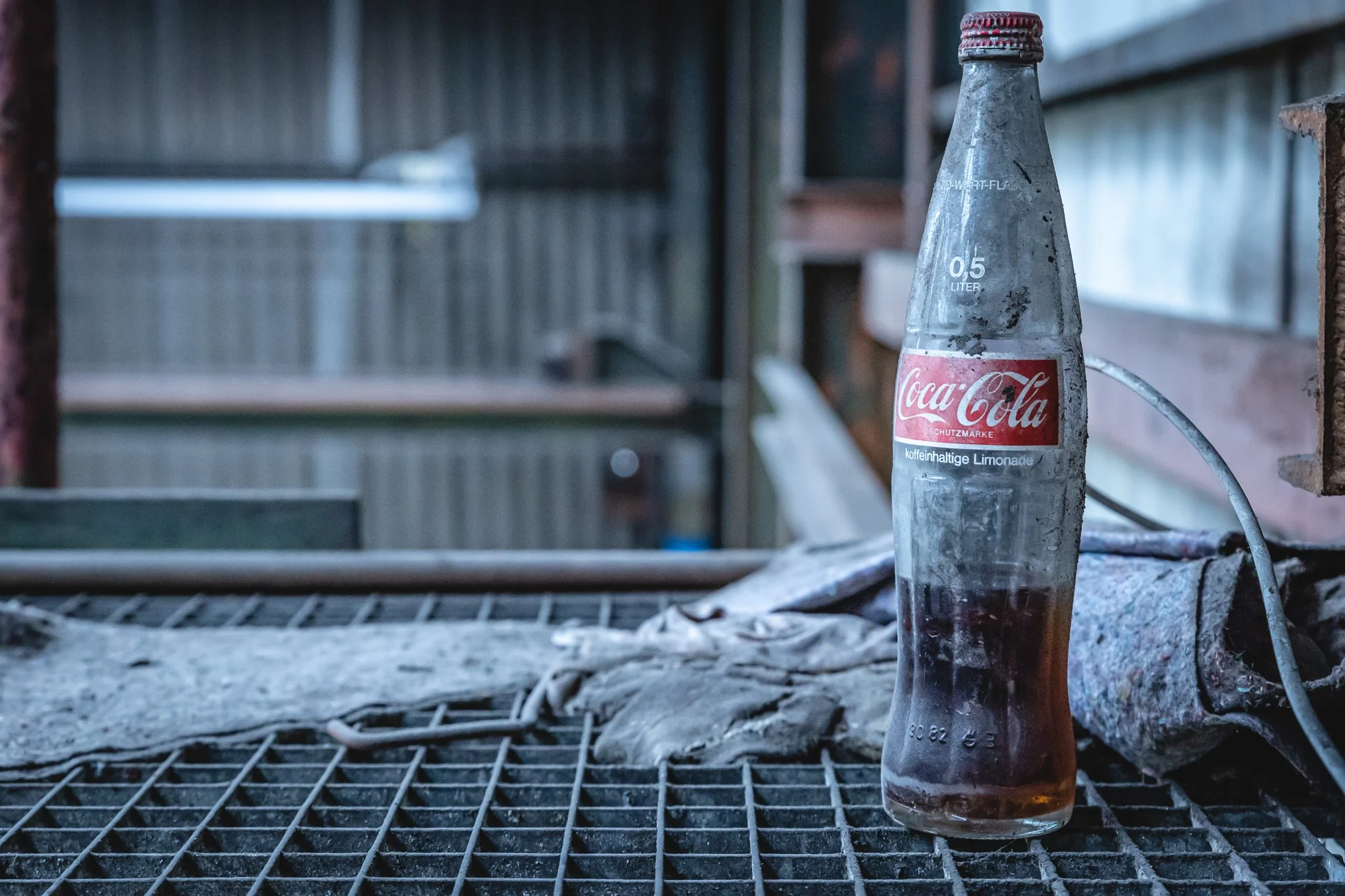 Eine leere Coca-Cola-Glasflasche mit rotem Verschluss und Etikett, die auf einem verrosteten Metallgitter in einer Außen- oder Industrieumgebung steht.