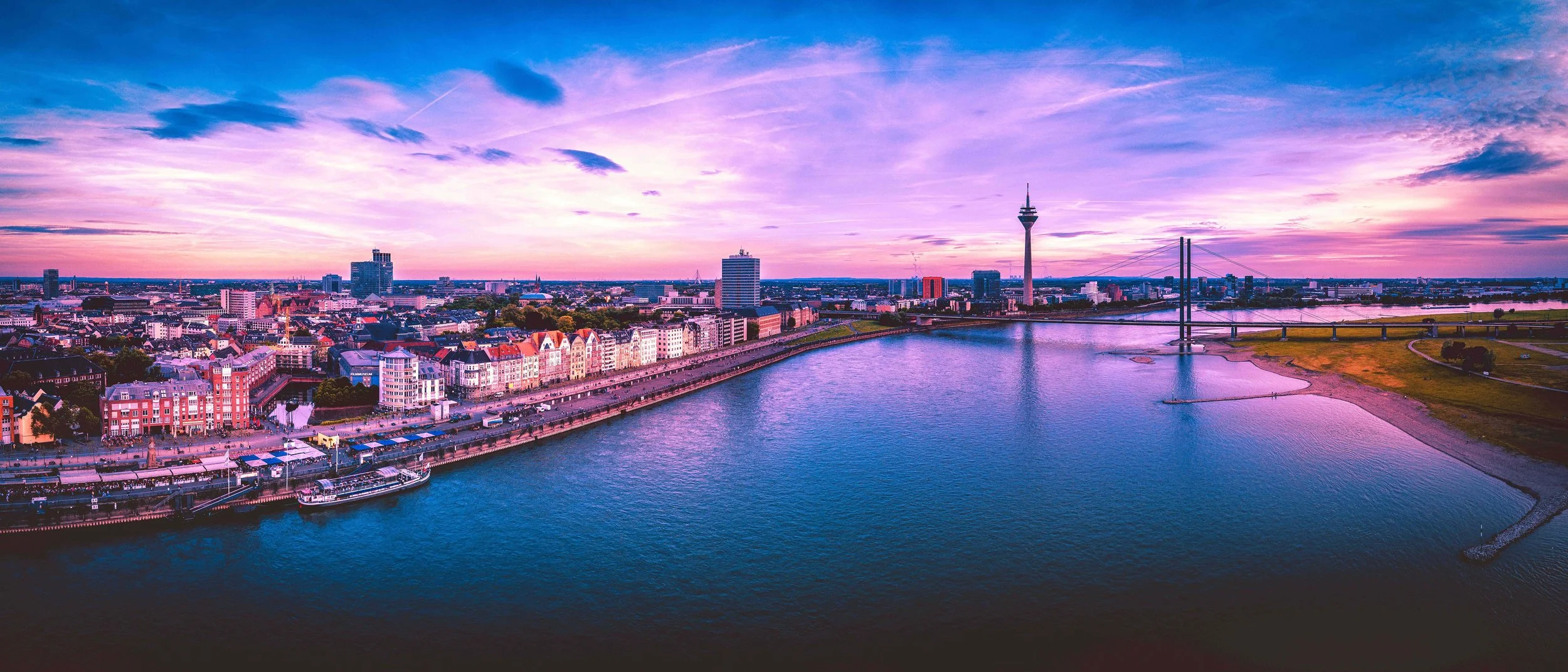 Stadtansicht von Düsseldorf mit Rheinufer, bunten Gebäuden, Fernsehturm und Hohenzollernbrücke bei Sonnenuntergang.