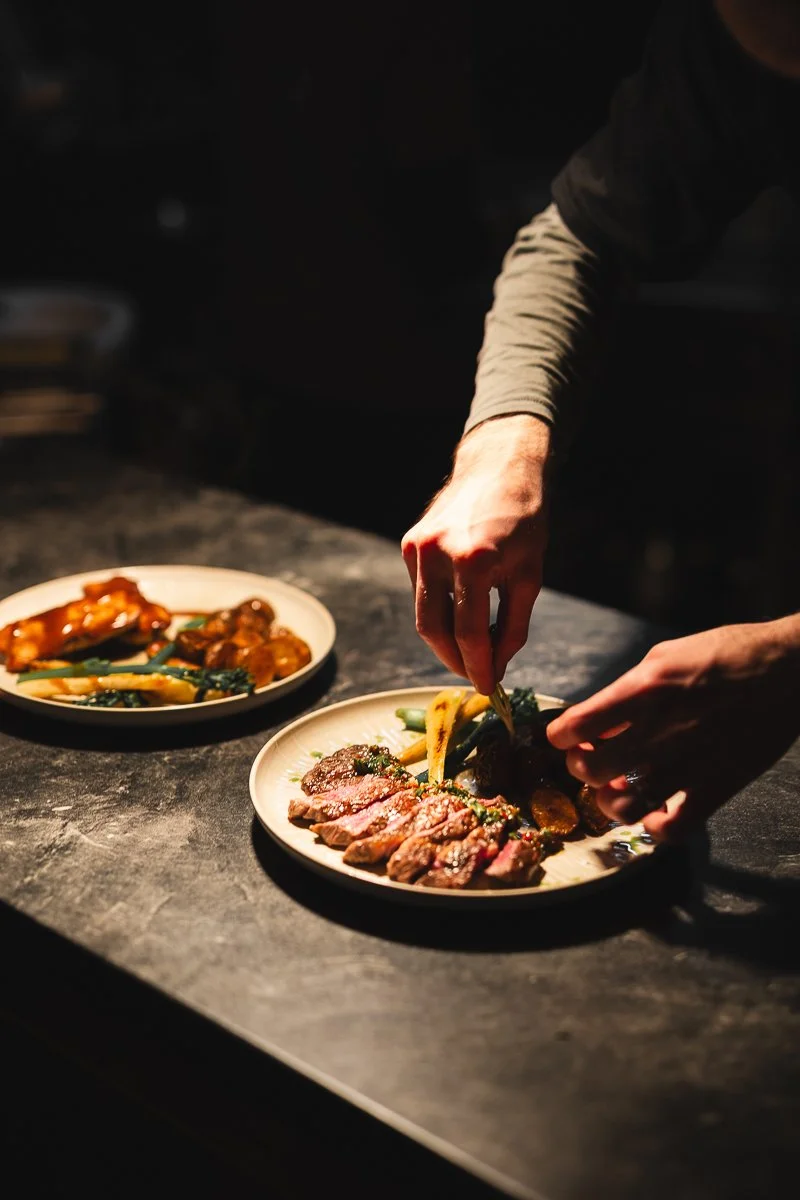 Ein Koch bereitet in einer schummrig beleuchteten Küche einen Teller mit Steakscheiben und Gemüse zu.