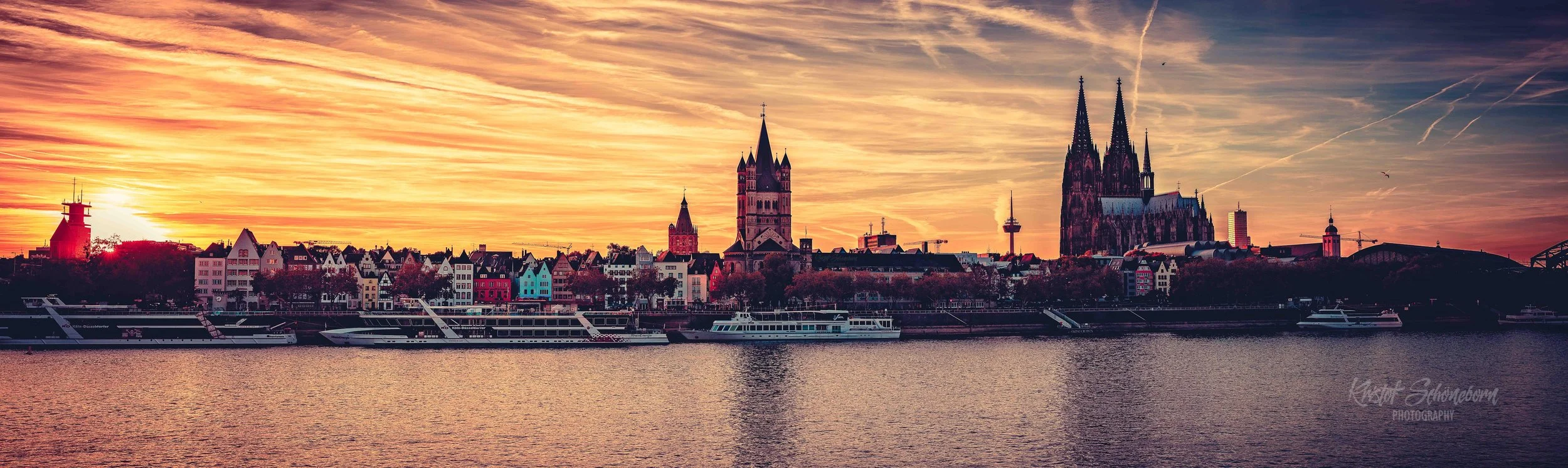 Panorama vom Sonnenuntergang über Köln mit Blick auf den Rhein, mit bunten Häusern, einer Kathedrale (Dom) und einem Fernsehturm, sowie Booten auf dem Fluss.
