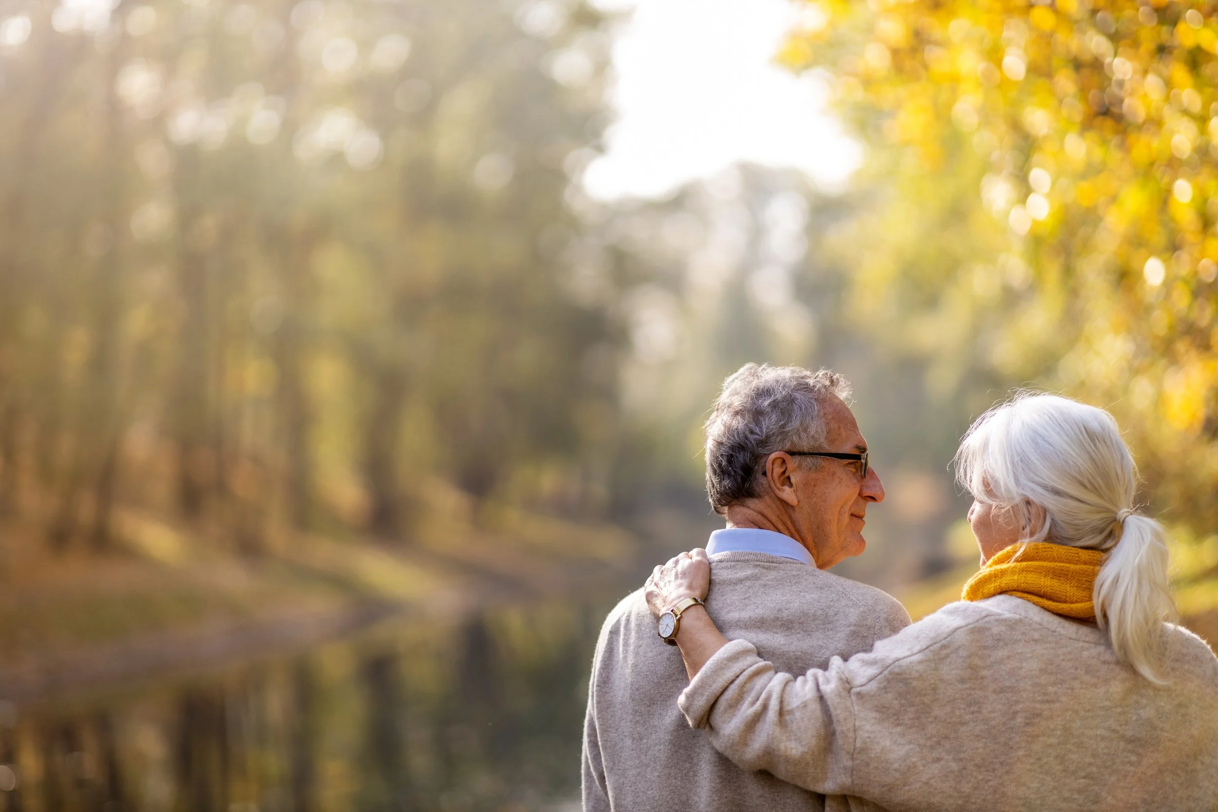 Older man and woman embracing outdoors during fall with colorful blurred trees in the background.