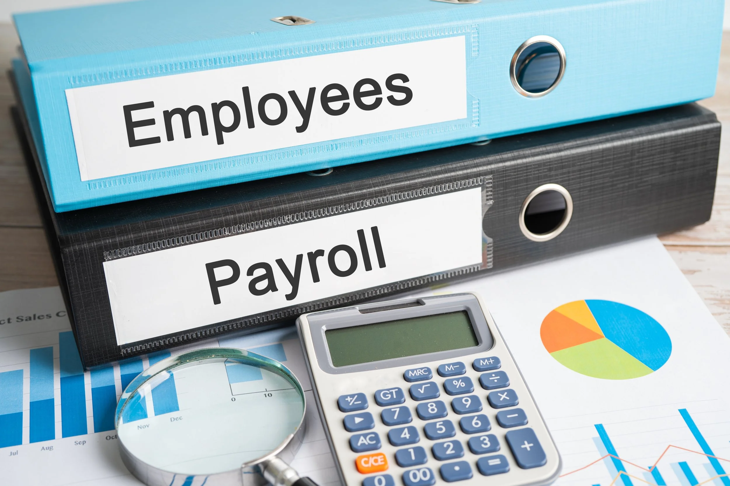 Blue and black binders labeled 'Employees' and 'Payroll' on a desk with a calculator, glasses, a pie chart, and financial reports.