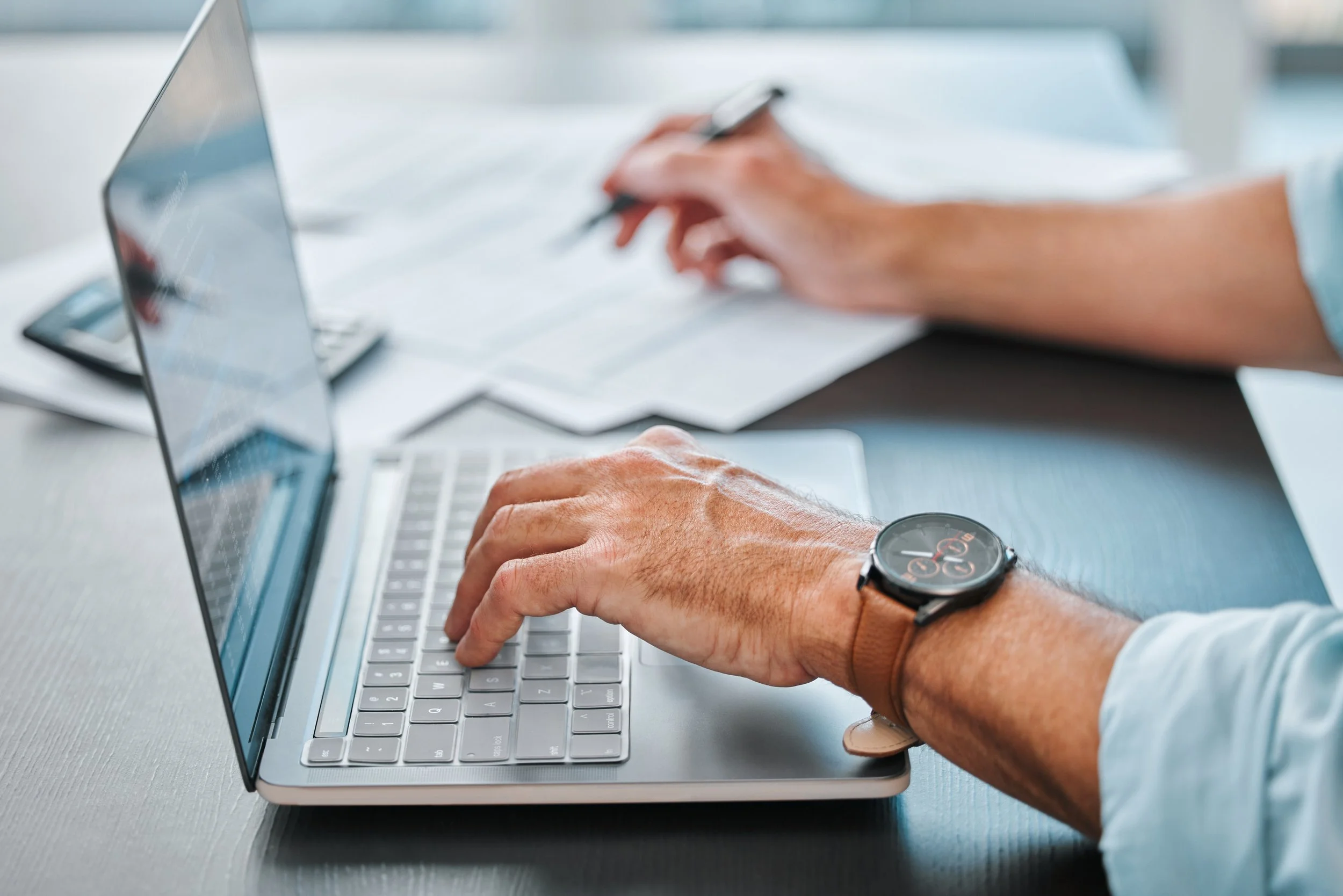 Close-up of a person's hands typing on a silver laptop keyboard, wearing a smartwatch with a round face.