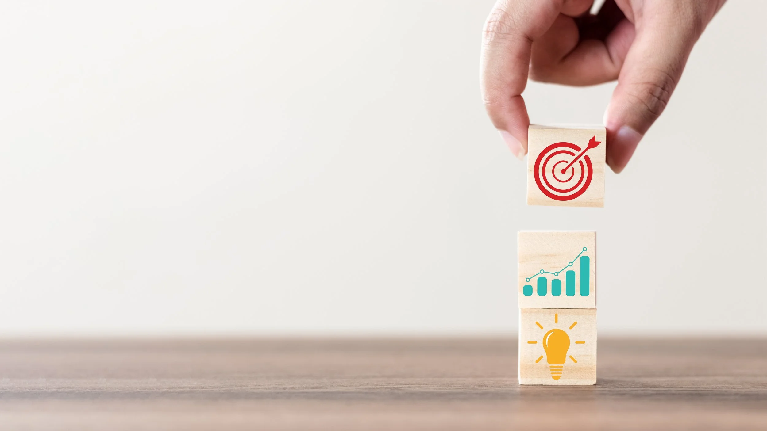 Hand stacking wooden blocks with icons: a target with an arrow, a bar chart with an upward trend, and a lightbulb.