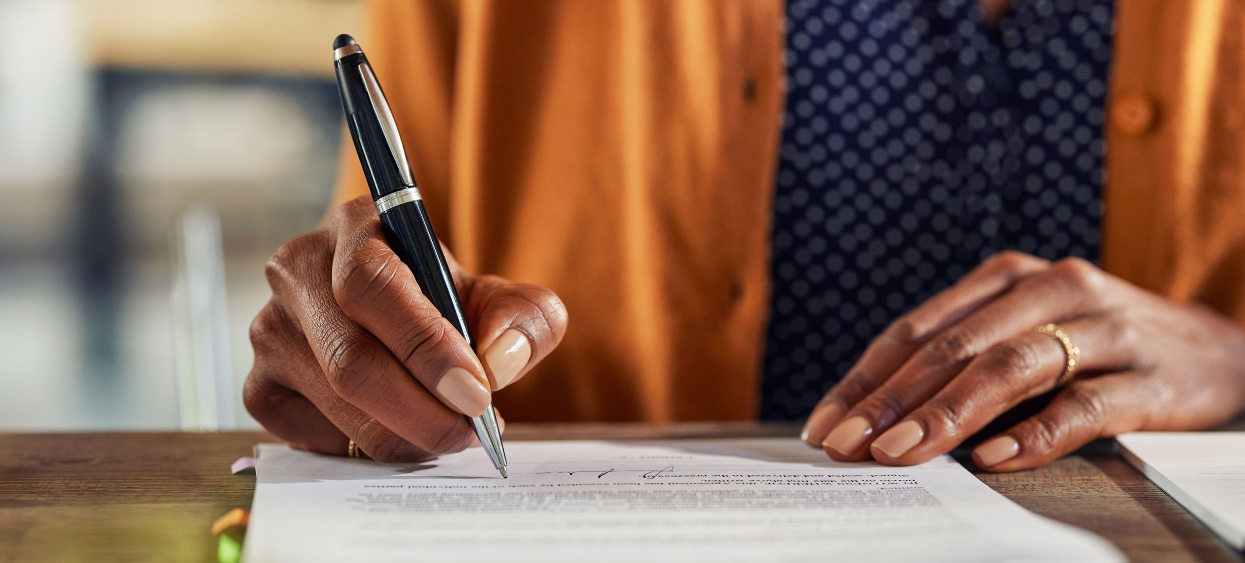 A person with dark skin is writing on a document with a black pen, sitting at a wooden desk. The person is wearing a brown jacket and a dark blue shirt with small white polka dots, along with a gold ring on the right hand.